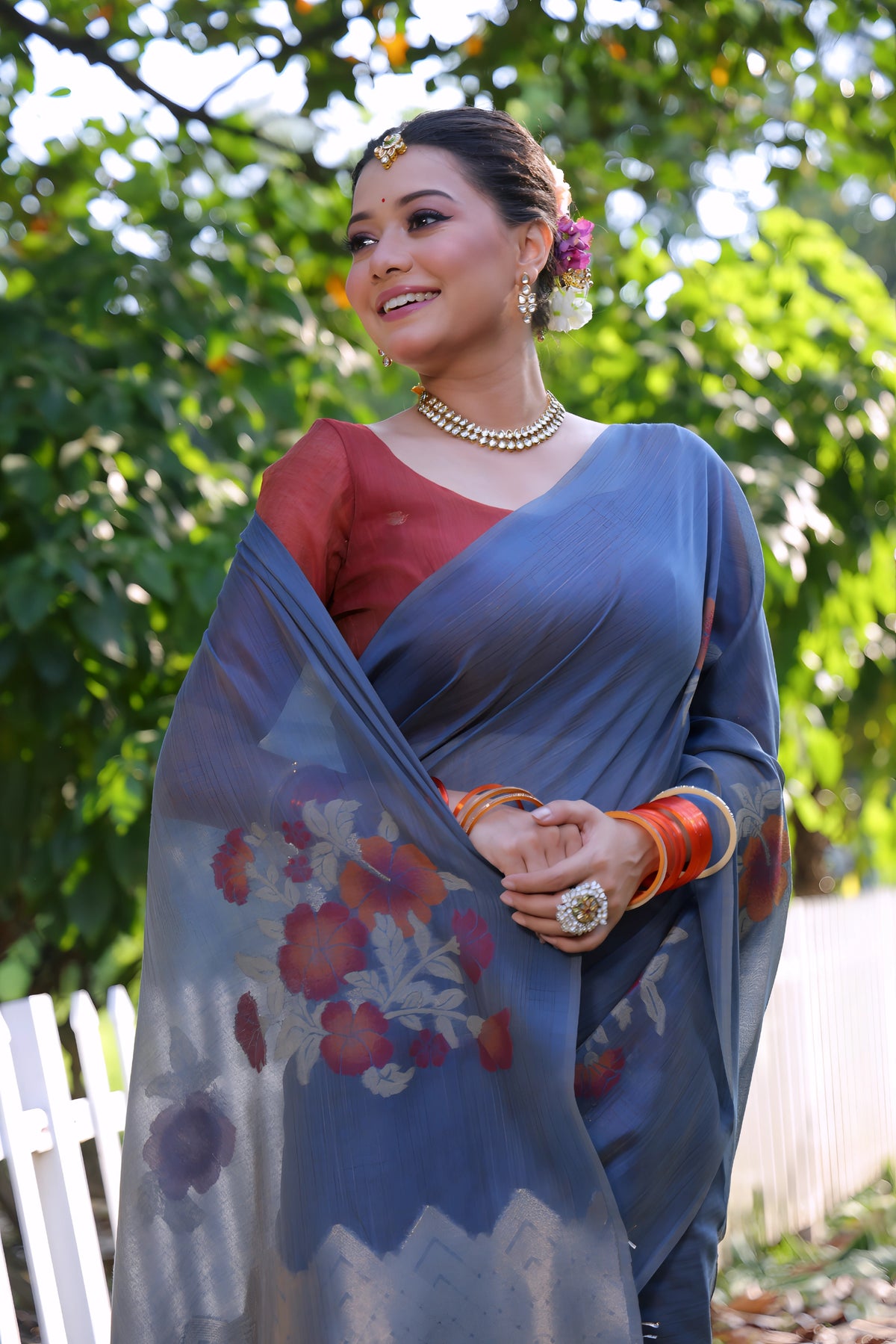 A woman models an elegant Slate Blue Soft Muga Cotton Saree. The pallu features large, colorful Resham floral weaving in red and orange. She wears a contrasting terracotta red blouse and is accessorized with a pearl choker necklace and vibrant orange bangles, posing outdoors by a white fence.