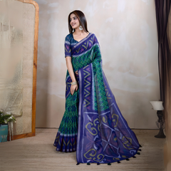 Woman wearing a blue and green handloom silk saree featuring bold geometric patterns, zigzag lines, and diamond motifs, paired with a matching blue blouse. She is styled with silver jewelry and bangles, posing gracefully in a contemporary studio setting with a neutral wall, a cane partition, a decorative lamp, and minimal furnishings for an elegant traditional look.