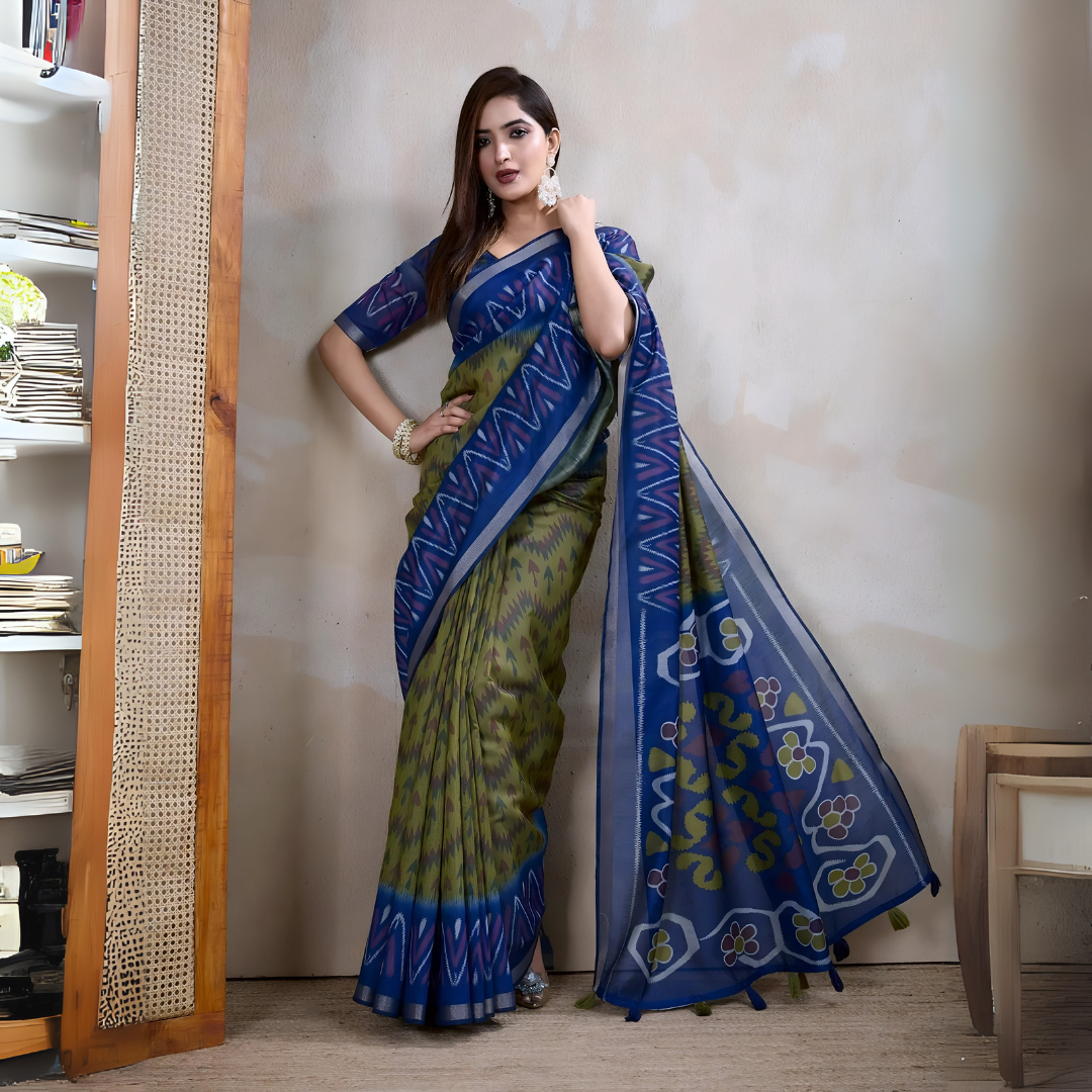 Woman wearing a blue and green Ikat silk saree featuring a geometric zigzag pattern and a striking blue pallu with floral and abstract motifs, paired with a matching blouse and traditional silver jewelry. She stands gracefully in a contemporary studio with a textured neutral wall, a woven cane screen, wooden furniture, and a bookshelf, creating an elegant and modern aesthetic.