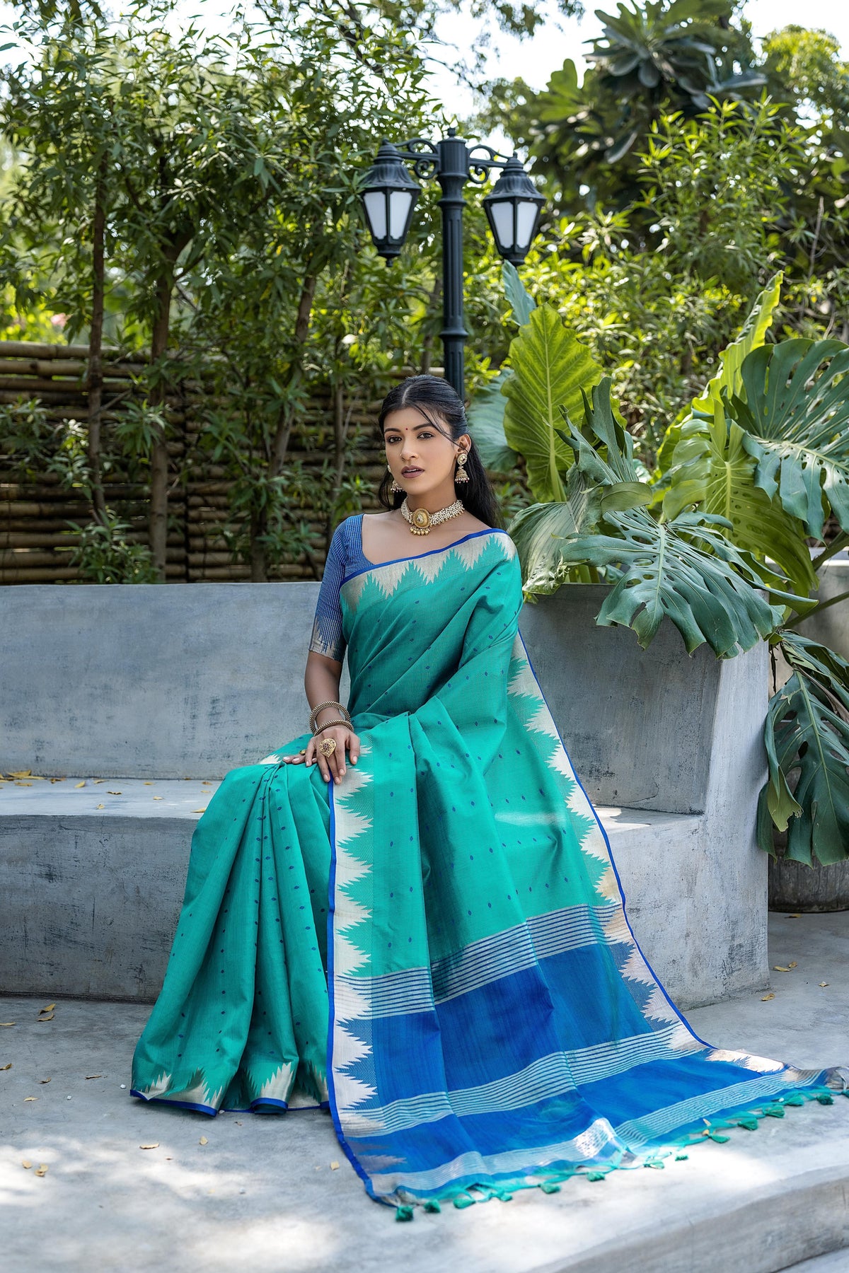 A woman models a striking Teal Blue Tussar Silk Saree with small woven motifs. The saree has a beige/silver Zari temple border and a pallu featuring wide bands of deep royal blue and Zari stripes. She wears a printed blue blouse and heavy gold Kundan jewelry.