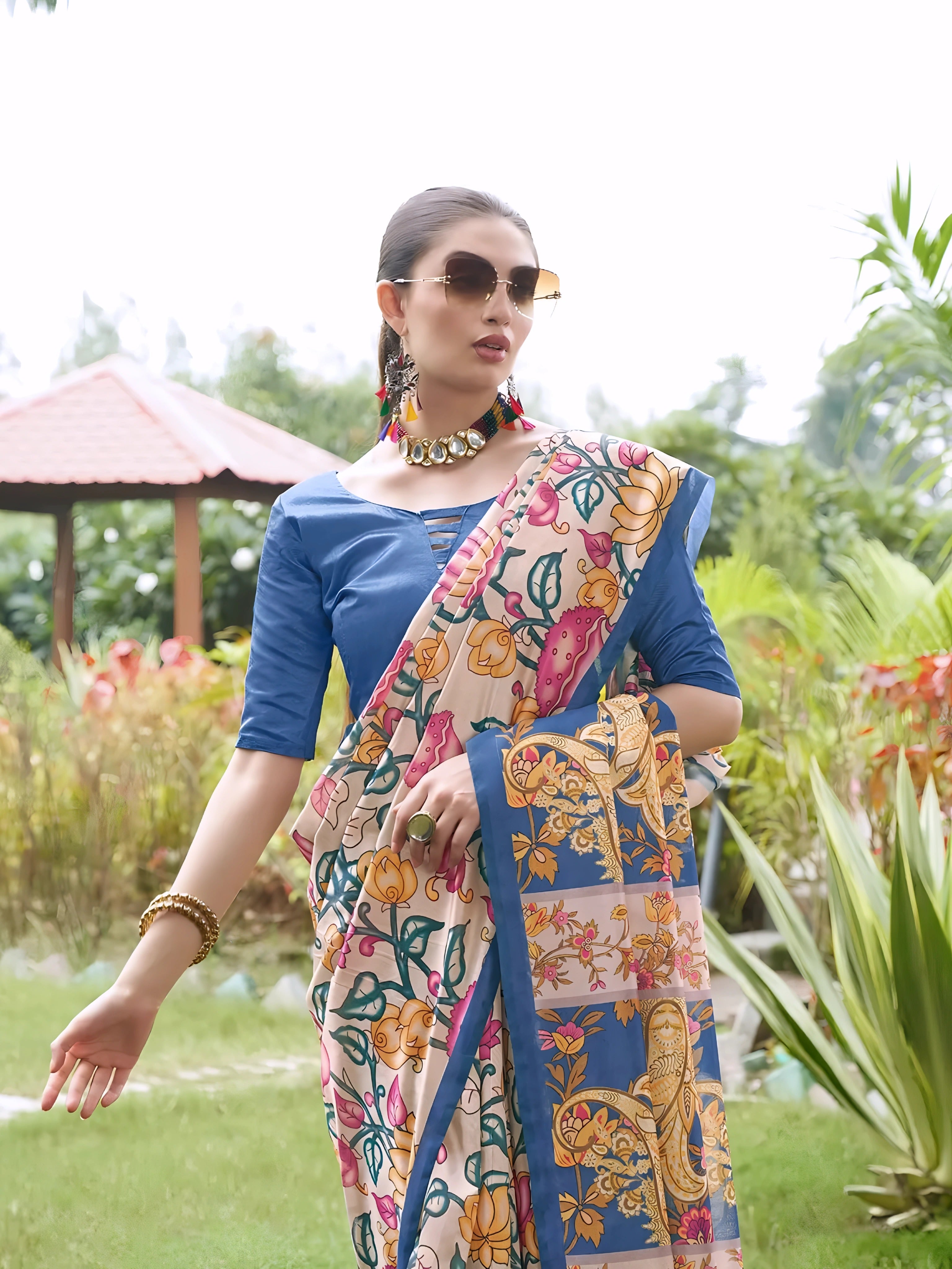 A woman models an elegant Beige Soft Tussar Silk Saree with a dense all-over print of multi-colored Kalamkari florals and vines (in pink, blue, and green). The saree has a wide, contrasting Deep Blue border and pallu with traditional print. She wears a matching blue blouse and a chunky beaded necklace, posing outdoors in a bright garden setting.