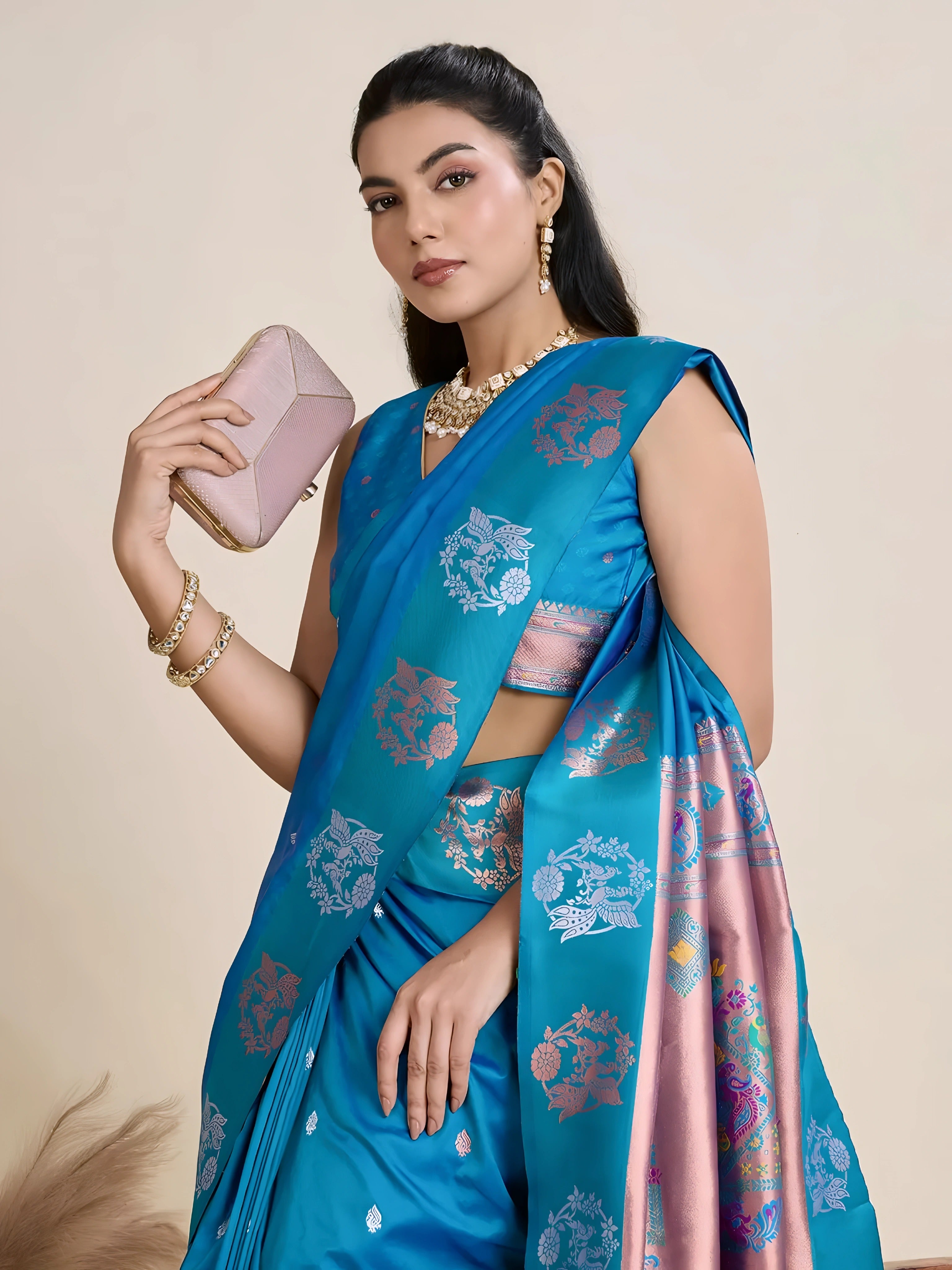 A woman models a luxurious Deep Blue Soft Silk Saree. The saree body features subtle metallic motifs. The pallu features a striking contrast panel in metallic rose gold/copper, richly woven with intricate blue and silver peacock and floral motifs. She wears a matching blue sleeveless blouse and heavy gold Kundan jewelry.