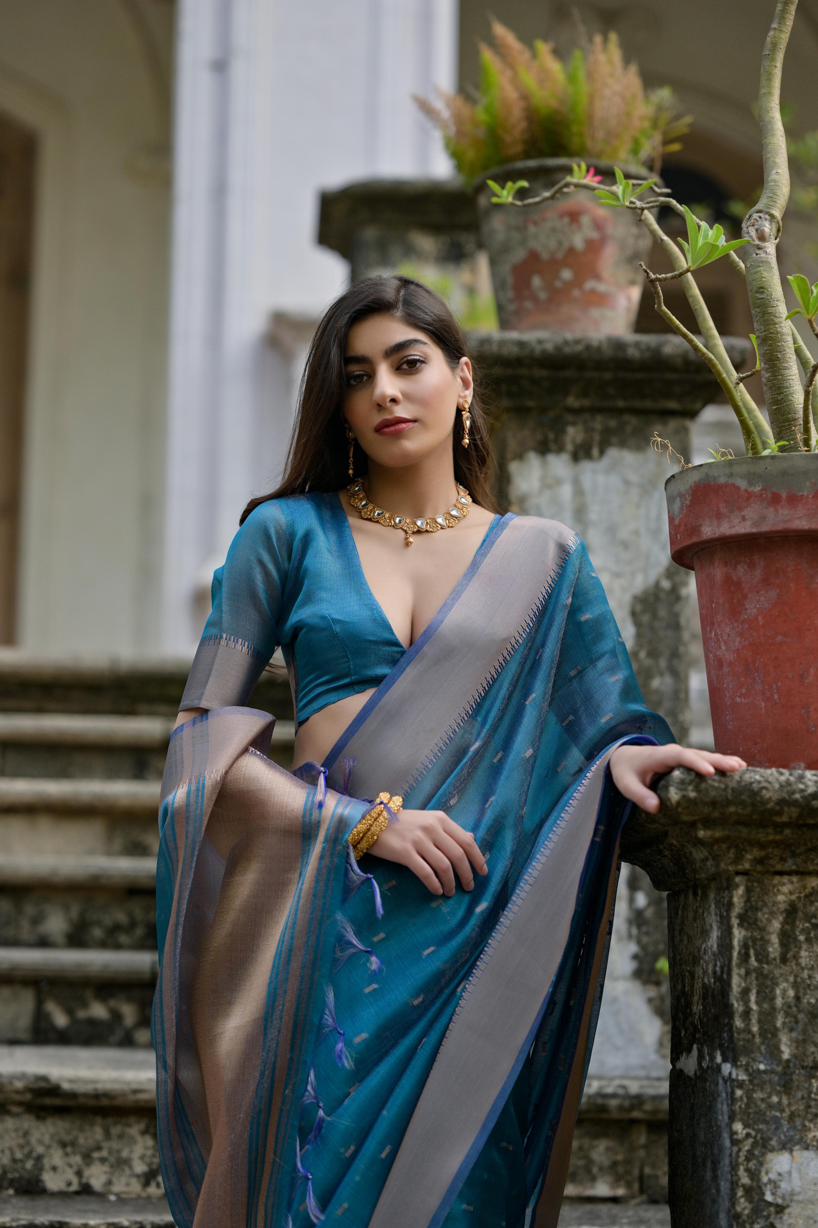 A woman models an elegant Deep Teal Soft Silk Saree. The saree features subtle metallic motifs on the body and a wide, contrasting matte silver/gray Zari woven border. She wears a dark grey short-sleeve blouse and a delicate gold choker necklace, posing gracefully on an ornate stone staircase.