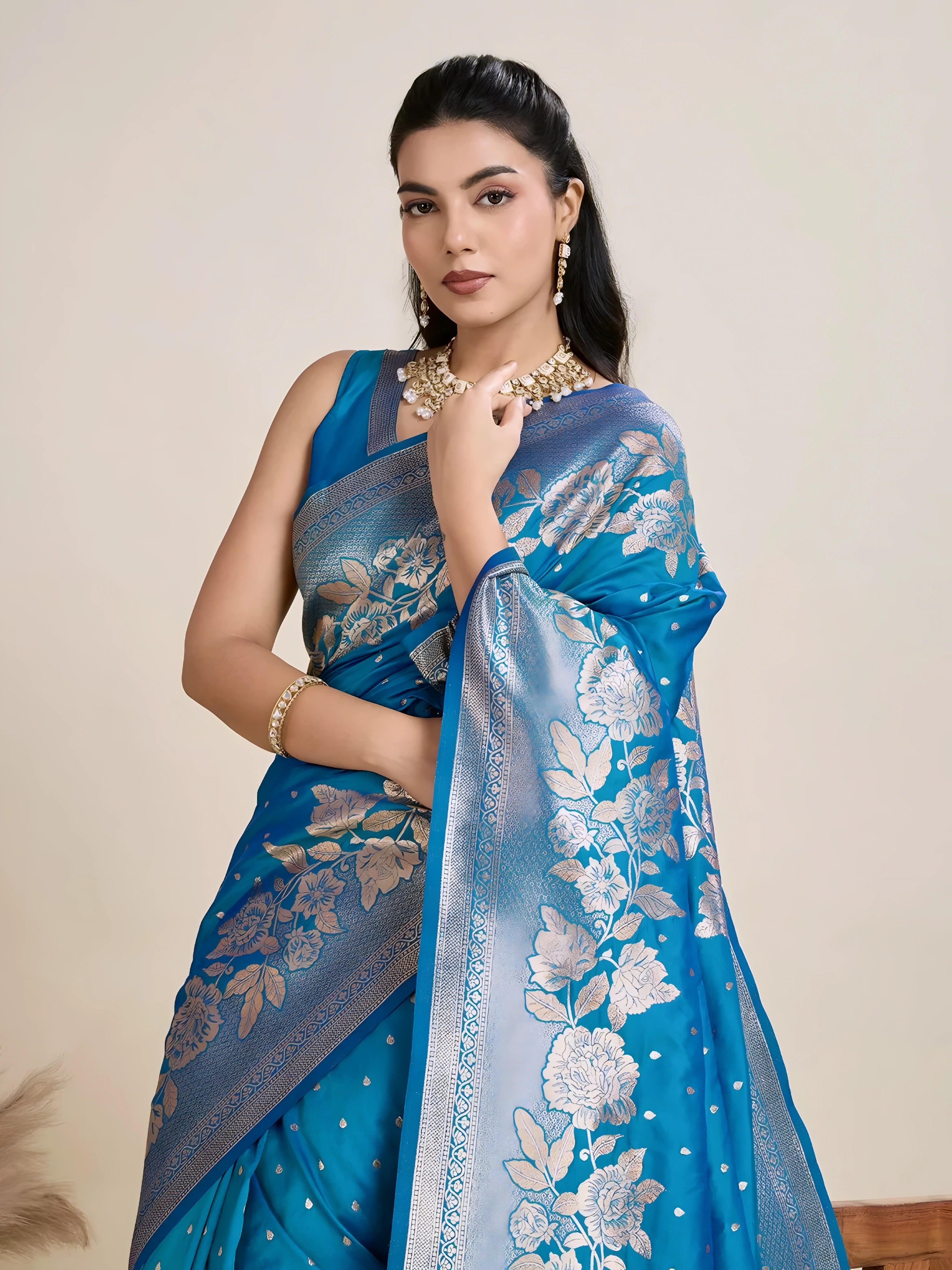 A woman models a luxurious Sea Blue Soft Kanjivaram Silk Saree. The saree features striking large floral and scattered motifs woven in metallic silver Zari on the body and wide pallu. She wears a matching blue sleeveless blouse and accessorizes with a heavy gold Kundan choker necklace and long earrings.