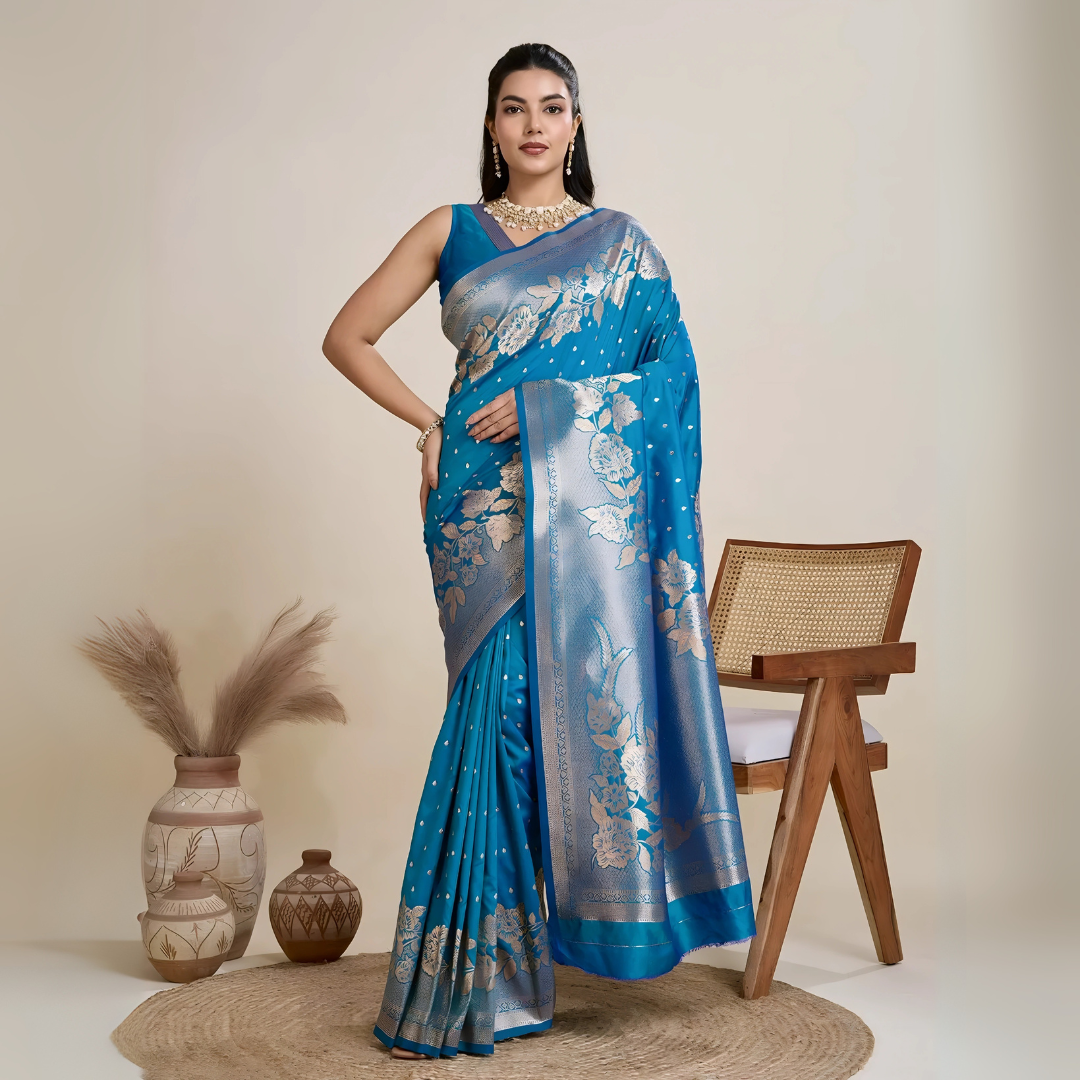 A woman models a luxurious Sea Blue Soft Kanjivaram Silk Saree. The saree features striking large floral and scattered motifs woven in metallic silver Zari on the body and wide pallu. She wears a matching blue sleeveless blouse and accessorizes with a heavy gold Kundan choker necklace and long earrings.