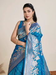 A woman models a luxurious Sea Blue Soft Kanjivaram Silk Saree. The saree features striking large floral and scattered motifs woven in metallic silver Zari on the body and wide pallu. She wears a matching blue sleeveless blouse and accessorizes with a heavy gold Kundan choker necklace and long earrings.