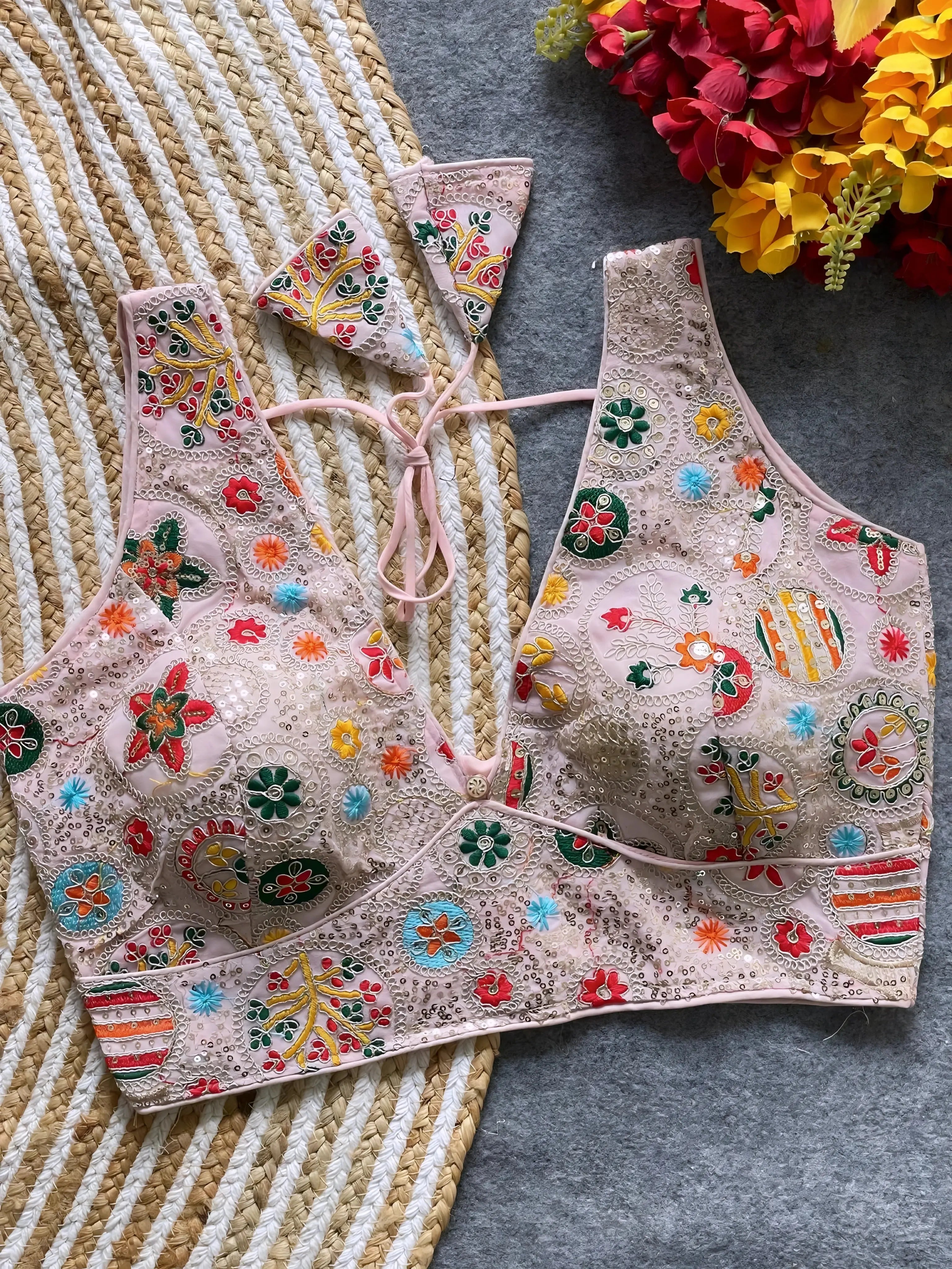 Traditional blush pink blouse featuring vibrant multicolor thread embroidery and mirror work in circular floral and geometric patterns, with sequin accents and a deep V-neck tie-back detail, displayed flat on a textured mat with artificial red and yellow flowers in the corner.