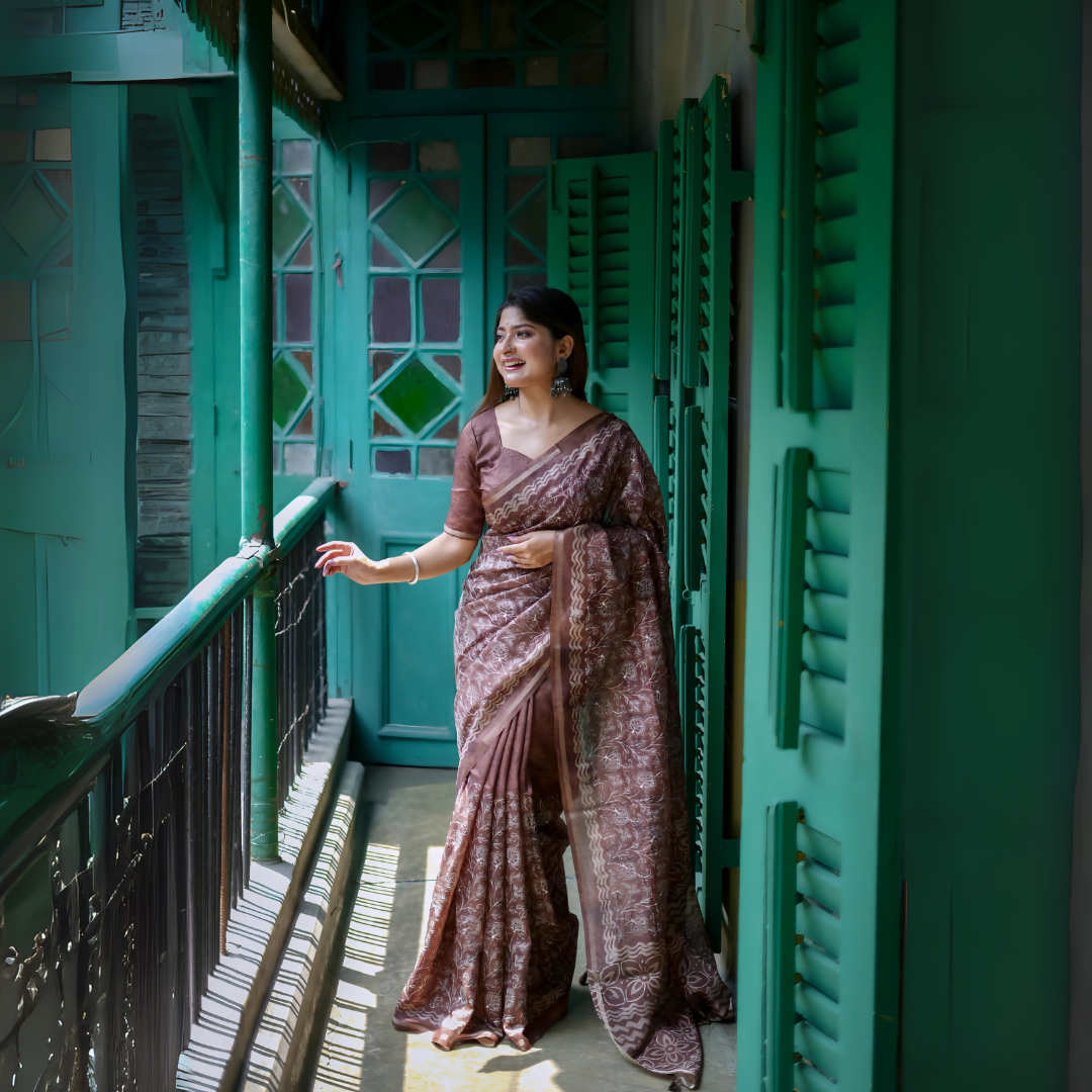 A woman models an elegant Dusty Brown Tussar Silk Saree. The saree features dense all-over floral embroidery/weaving in a subtle, tone-on-tone thread. She wears a matching brown short-sleeve blouse and accessorizes with large oxidized silver jhumka earrings and bangles, posing by vibrant teal green shutters.