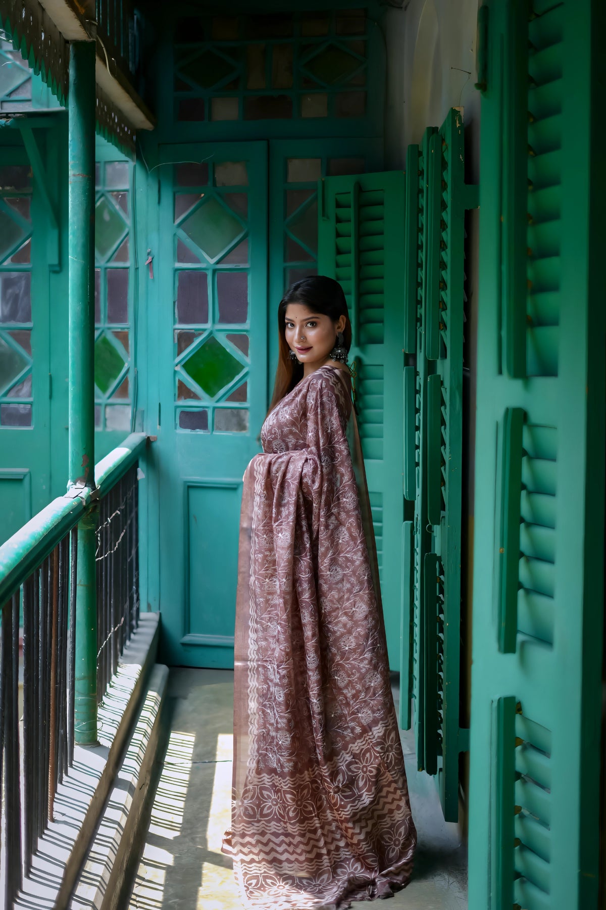 A woman models an elegant Dusty Brown Tussar Silk Saree. The saree features dense all-over floral embroidery/weaving in a subtle, tone-on-tone thread. She wears a matching brown short-sleeve blouse and accessorizes with large oxidized silver jhumka earrings and bangles, posing by vibrant teal green shutters.