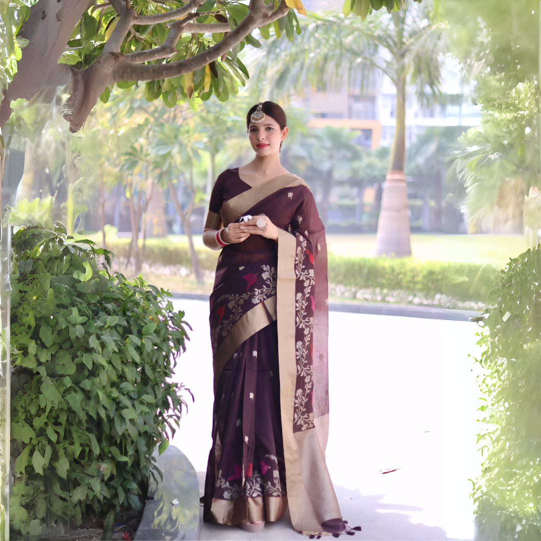 A woman models an elegant Deep Plum Linen Silk Saree. The saree features delicate woven floral and bird motifs and a wide, contrasting pale gold Zari border. She wears a matching plum blouse, contrasting red and white bangles, and a traditional gold maang tikka, posing outdoors.