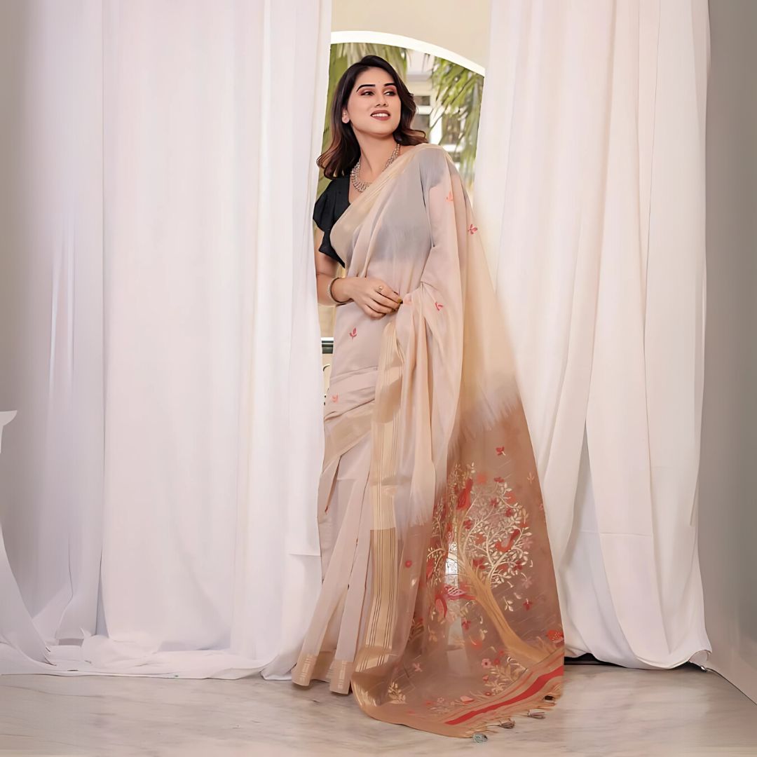 A woman models a Soft Muga Cotton Saree. The saree is light beige/cream, transitioning to a brown/rust-colored pallu featuring an elaborate, colorful Resham and Zari woven design of a tree and birds. She wears a black ruffled blouse and an oxidized silver necklace.