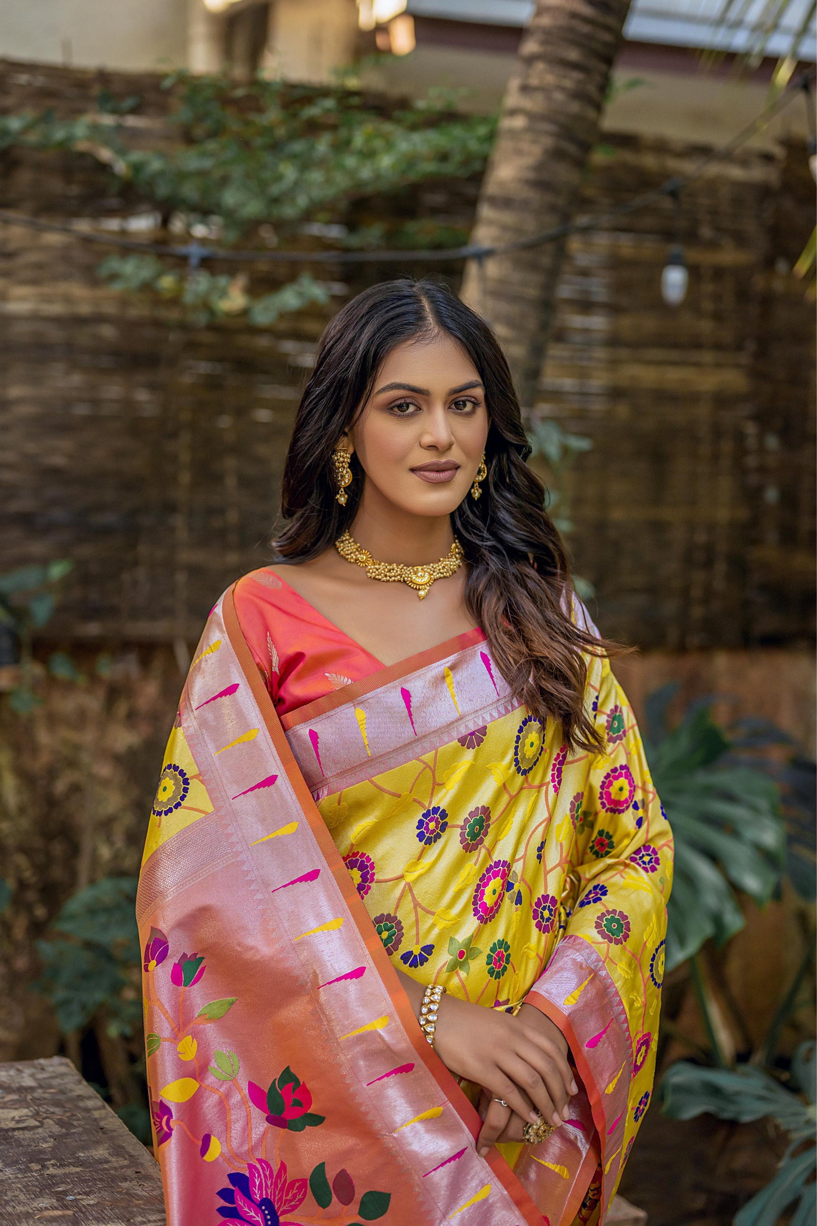 Close-up of woman in brown yellow zari work saree with intricate floral pallu, styled with traditional gold jewelry in an outdoor setting
