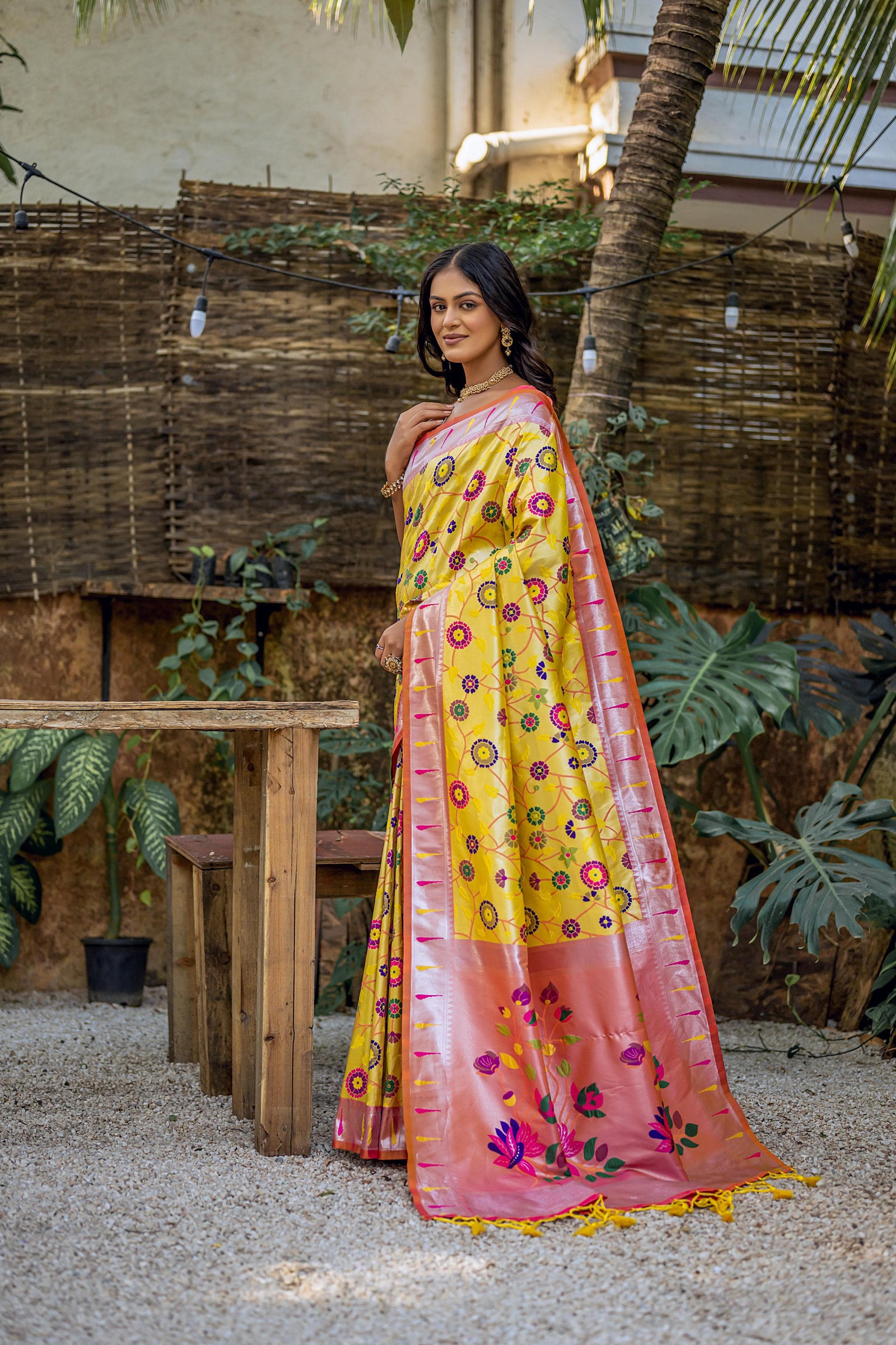 Woman wearing brown yellow zari work saree with peach floral pallu, posing in a garden setting for a traditional and elegant look