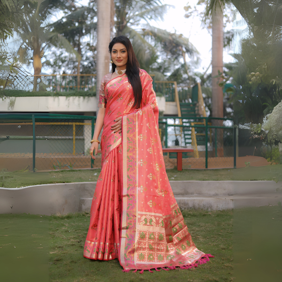 Woman wearing a coral pink Banarasi silk saree adorned with traditional gold zari motifs and a broad multicolor pallu featuring intricate temple and floral patterns, paired with a matching short sleeve blouse and gold jewelry. She stands gracefully on a lush green lawn with landscaped garden elements, outdoor stairs, and tropical trees in the background, creating an elegant