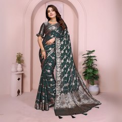 Woman wearing a dark green Banarasi saree with intricate silver floral motifs, a rich ethnic border, and elegant pallu design. The saree is paired with a matching silver blouse featuring short sleeves and delicate detailing. Styled with traditional jewelry and bangles, she poses gracefully in front of a pastel arched backdrop with minimalist decor, including a white pedestal and a potted green plant.
