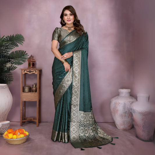 Woman wearing a dark green Banarasi silk saree featuring an ornate golden zari border and intricate circular woven motifs, paired with a matching green blouse and traditional jewelry. She poses gracefully in a modern studio setting with large white clay pots, a wooden stand adorned with brass decor, leafy green plants, and a bowl of fresh marigolds on the floor.