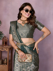 Woman wearing a dark green Banarasi silk saree featuring an ornate golden zari border and intricate circular woven motifs, paired with a matching green blouse and traditional jewelry. She poses gracefully in a modern studio setting with large white clay pots, a wooden stand adorned with brass decor, leafy green plants, and a bowl of fresh marigolds on the floor.