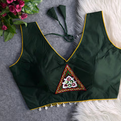 Dark green padded blouse with yellow piping, featuring a central embroidered triangle motif with mirror work and shell detailing along the hem, displayed on a grey surface with artificial pink flowers and white fur in the background.