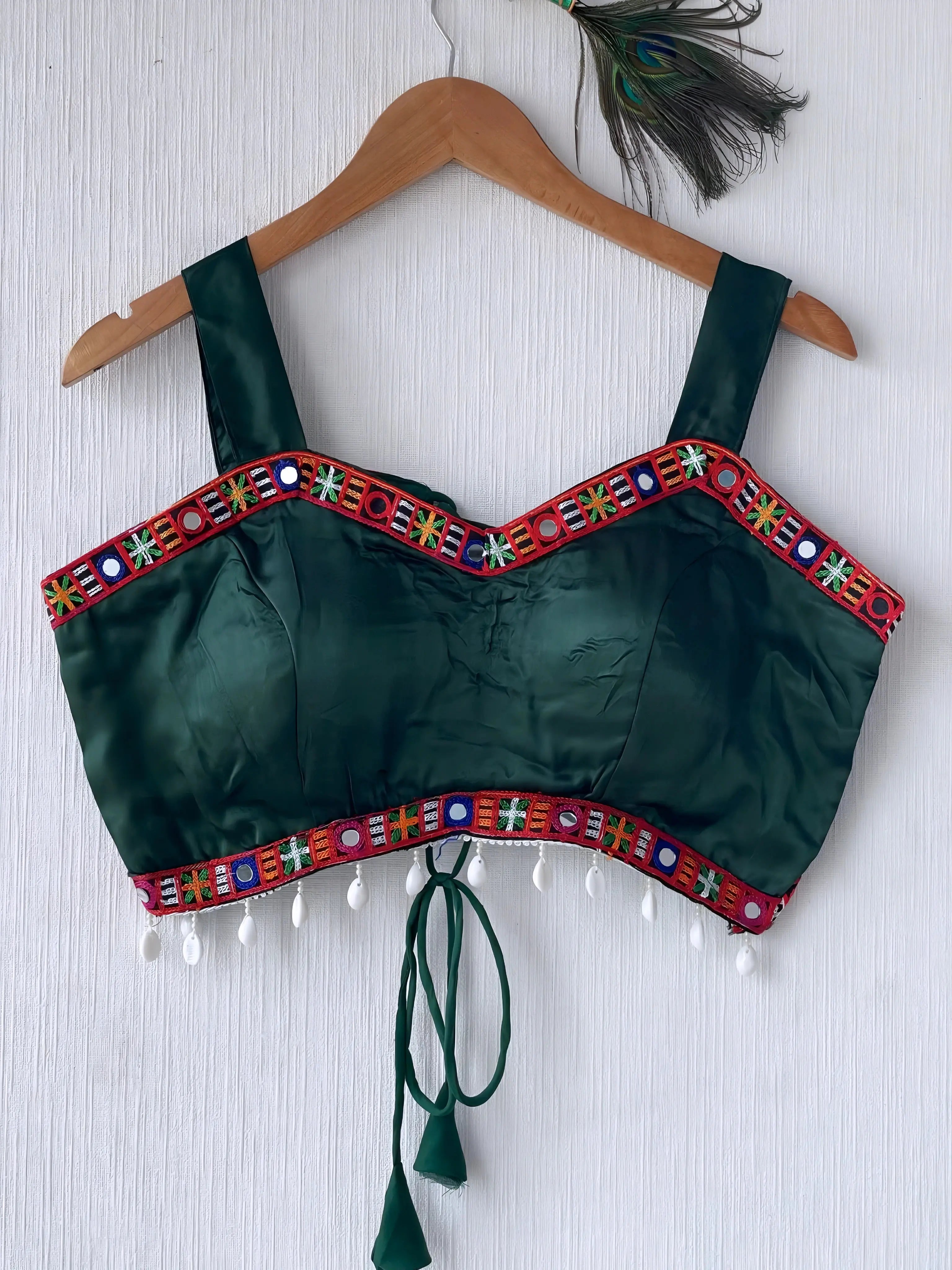 Dark green ethnic blouse featuring vibrant mirror work and multicolored embroidered border in red, blue, green, and white, embellished with pearl drop tassels and a tie-up at the back. Displayed on a wooden hanger against a textured off-white wall with peacock feathers for a festive, traditional look.