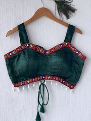 Dark green ethnic blouse featuring vibrant mirror work and multicolored embroidered border in red, blue, green, and white, embellished with pearl drop tassels and a tie-up at the back. Displayed on a wooden hanger against a textured off-white wall with peacock feathers for a festive, traditional look.