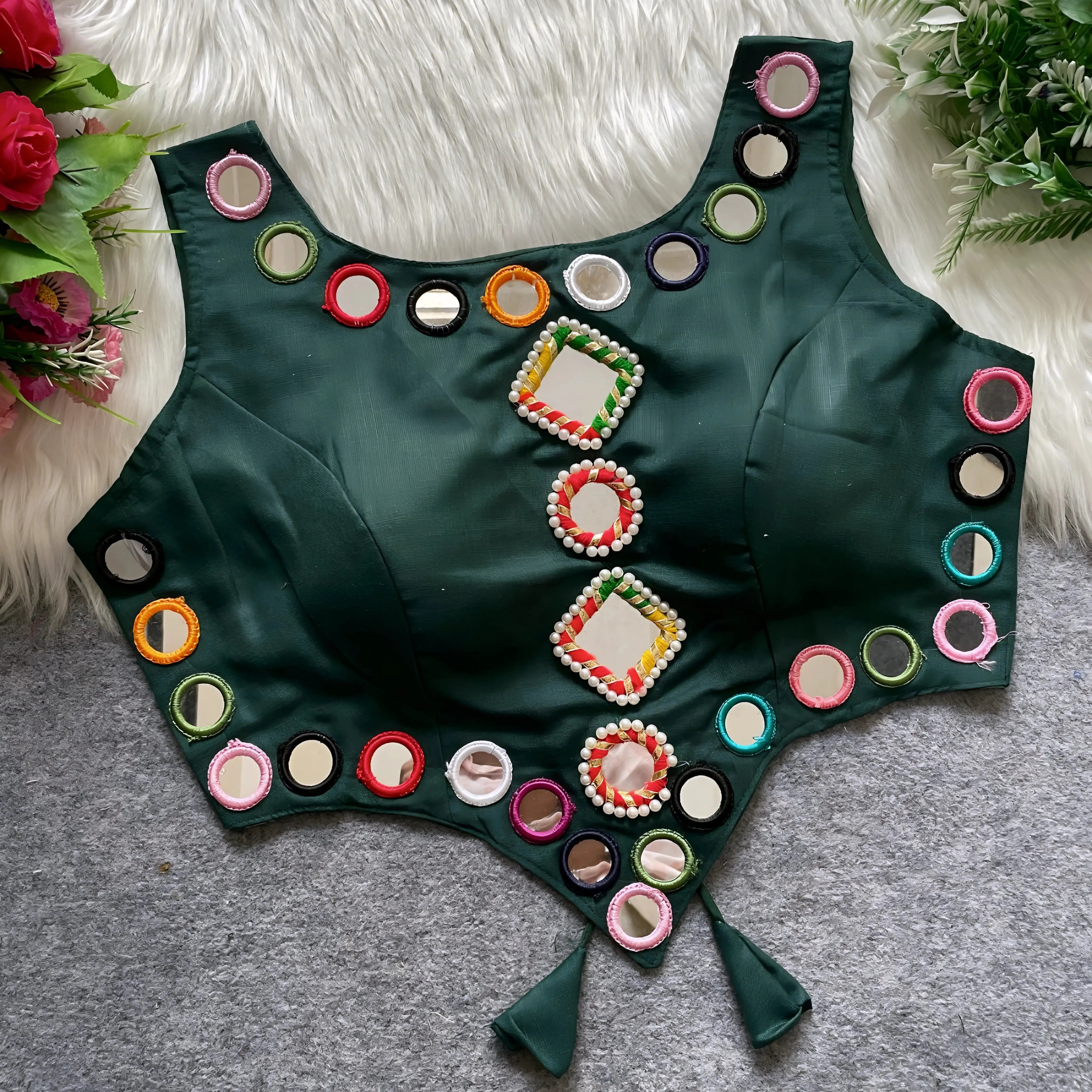  Dark green ethnic blouse featuring vibrant mirror work with multicolored embroidered borders and decorative diamond and round mirrors arranged in the center, adorned with pearl details and tie-up tassels at the bottom. Displayed flat on a grey surface with white faux fur and floral accents for a festive and traditional look.
