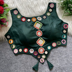  Dark green ethnic blouse featuring vibrant mirror work with multicolored embroidered borders and decorative diamond and round mirrors arranged in the center, adorned with pearl details and tie-up tassels at the bottom. Displayed flat on a grey surface with white faux fur and floral accents for a festive and traditional look.