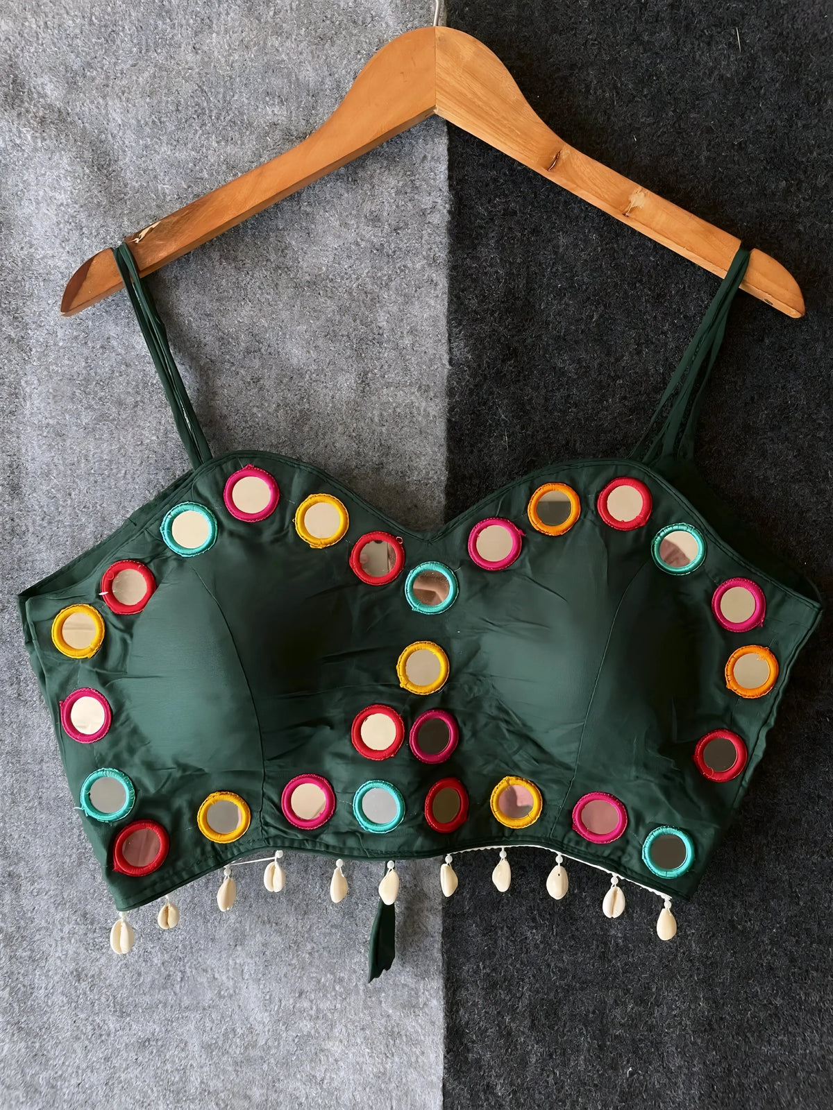 Dark green corset-style blouse embellished with vibrant round mirrors and multicolored embroidered borders, featuring thin shoulder straps and shell tassel drops along the hem. Displayed on a wooden hanger against a two-tone grey textured background, highlighting its festive and modern ethnic appeal.