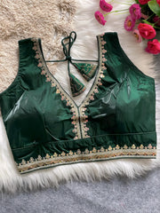Traditional dark green silk blouse featuring intricate gold and pastel embroidery along the neckline and hem, with decorative tassel tie at the back, displayed flat on a soft fur and textured mat background with artificial pink flowers in the corner.