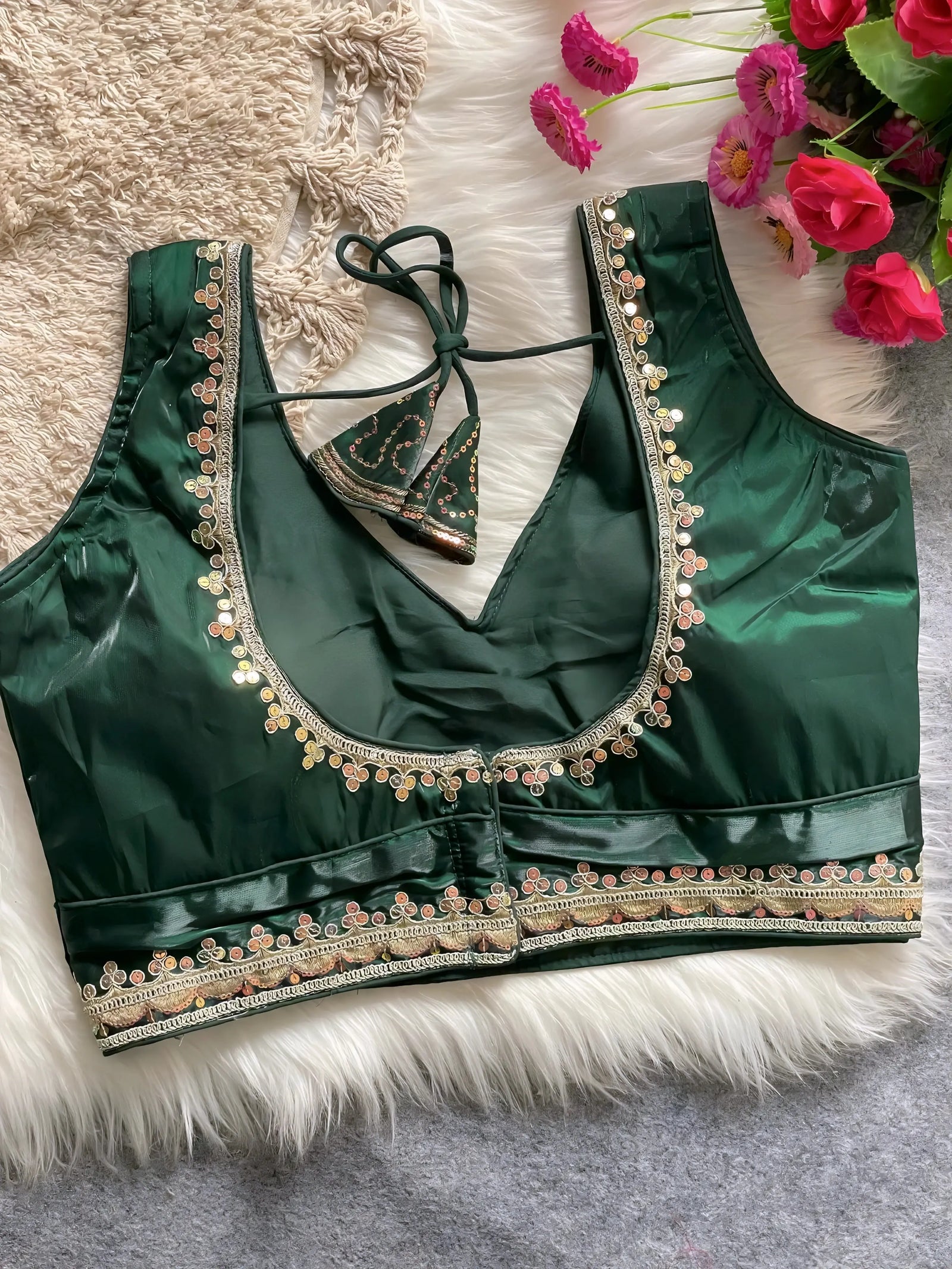 Traditional dark green silk blouse featuring intricate gold and pastel embroidery along the neckline and hem, with decorative tassel tie at the back, displayed flat on a soft fur and textured mat background with artificial pink flowers in the corner.