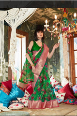 Woman in a dark green zari work saree with pink border standing indoors surrounded by colorful cushions and bohemian decor elements like macrame and a chandelier