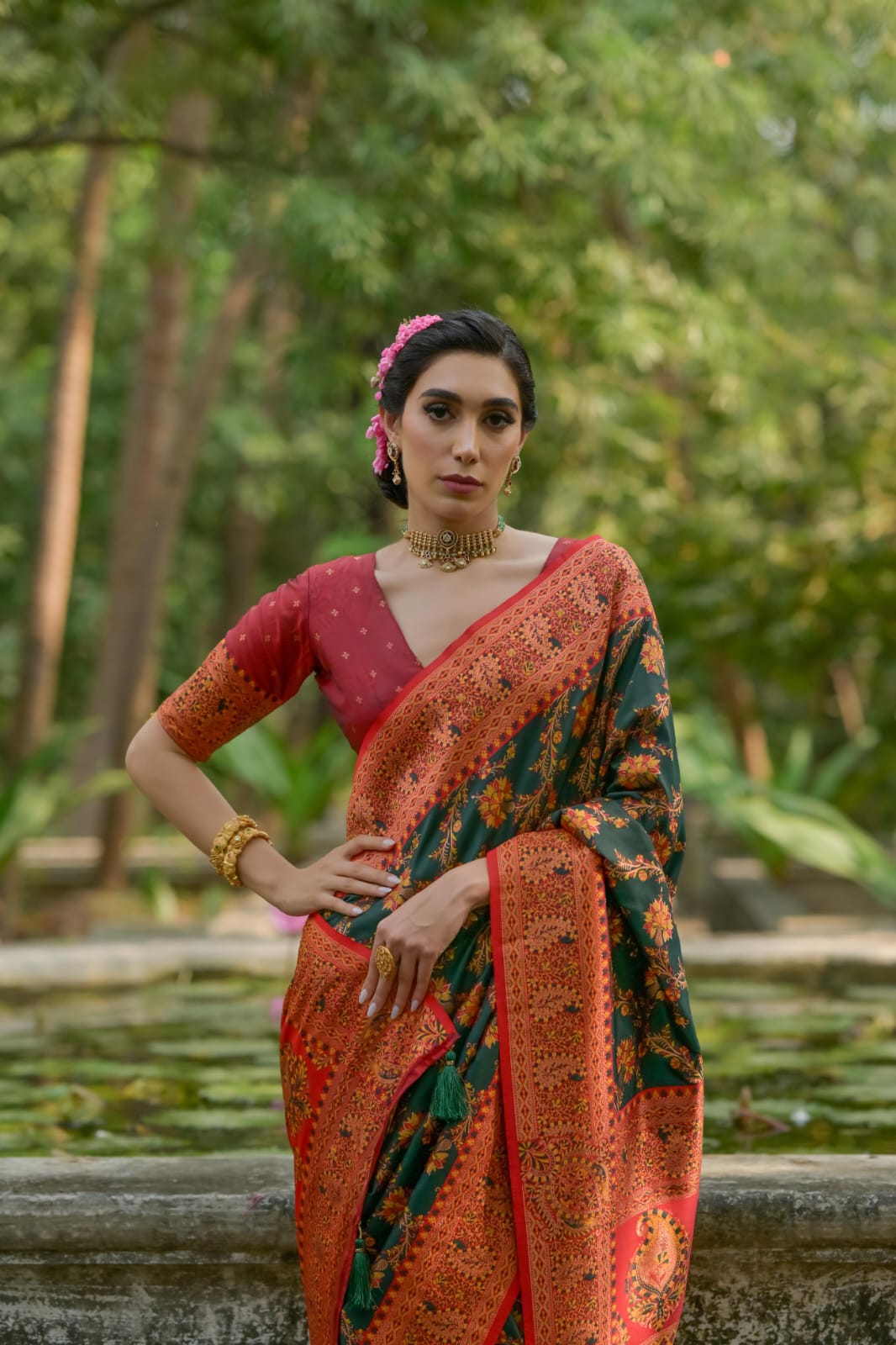 A person draped in a dark green saree featuring intricate zari work, posing gracefully in an outdoor setting with stone structures and lush greenery.