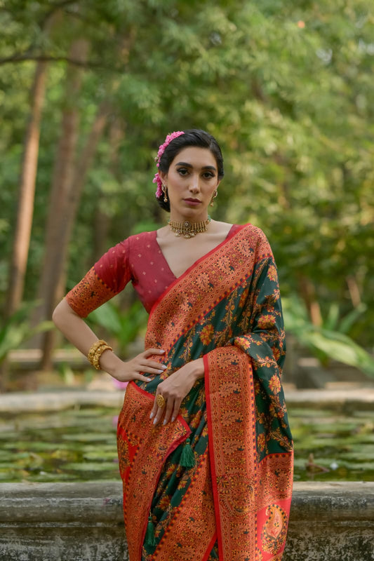 A person draped in a dark green saree featuring intricate zari work, posing gracefully in an outdoor setting with stone structures and lush greenery.