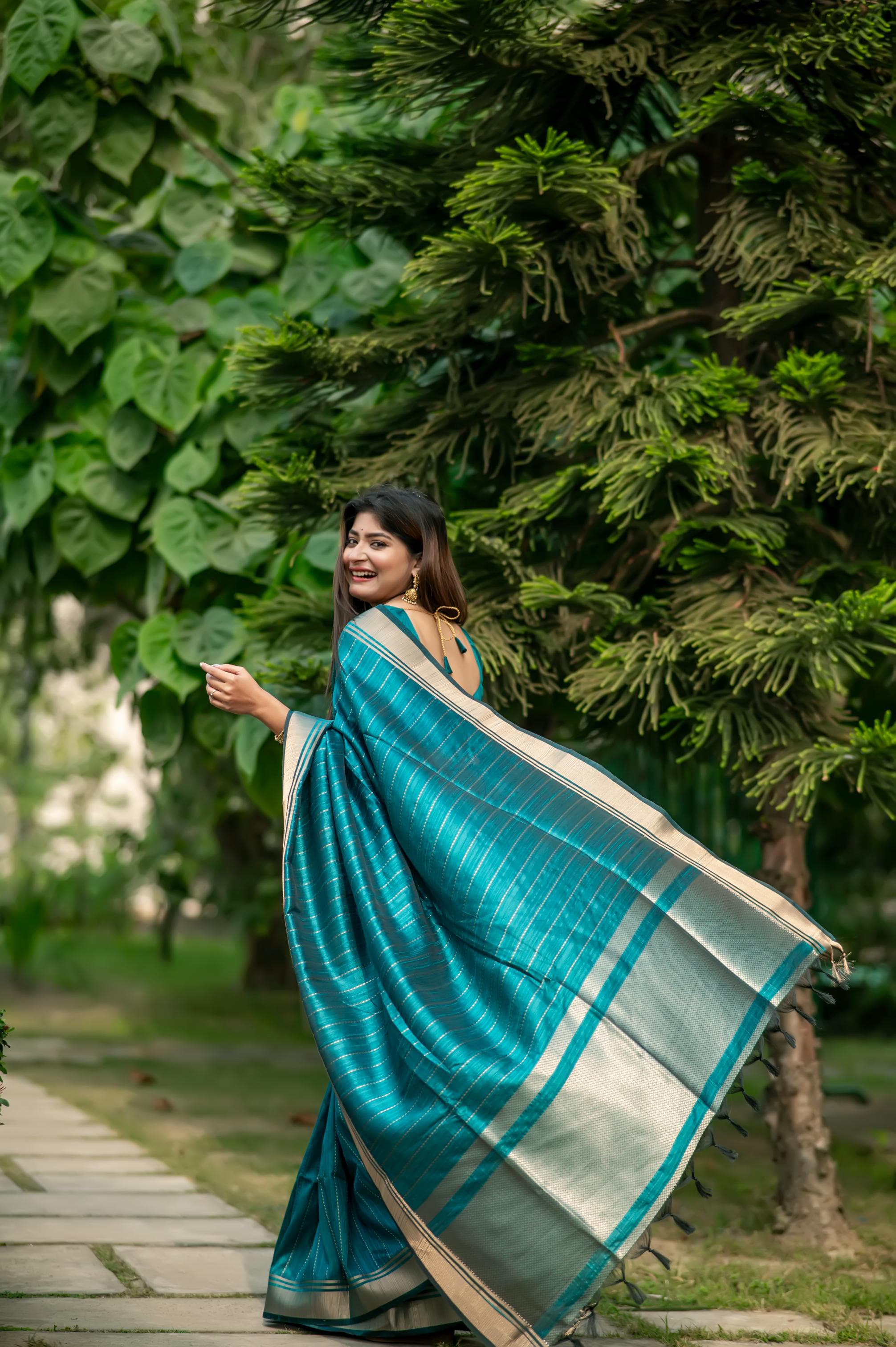 Woman in a dark turquoise saree with zari work posing gracefully on a garden pathway with lush green trees in the background