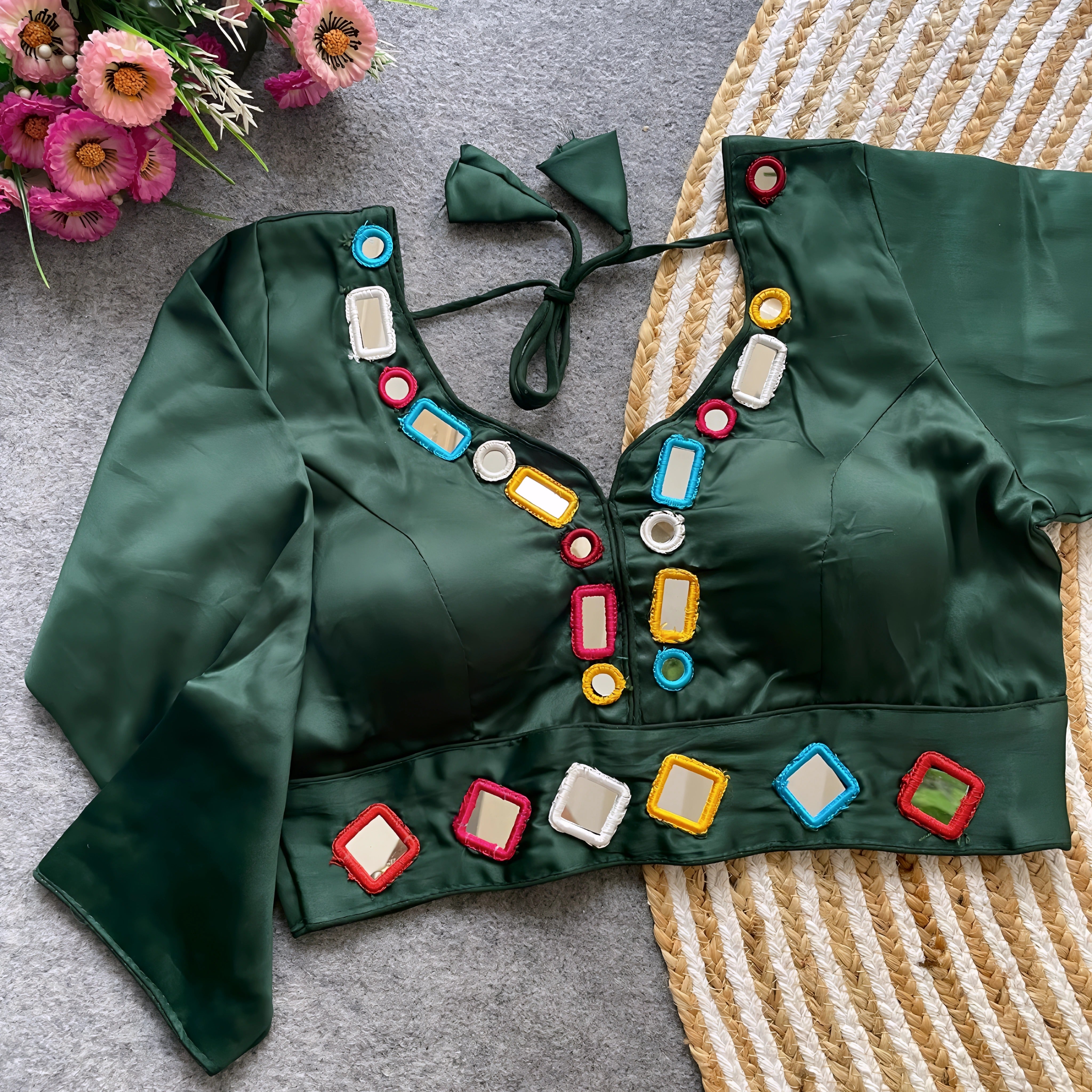 Dark green blouse embellished with square and round mirror work framed by multicolored embroidered borders, stylish back tie-up closure, and three-quarter sleeves. Displayed flat on a textured grey surface with a woven mat and fresh flowers, highlighting its festive and contemporary ethnic design.