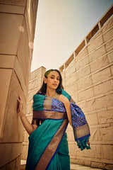 Elegant woman draped in deep green saree with rich zari work and intricate pallu design, posing against contemporary sandstone architecture