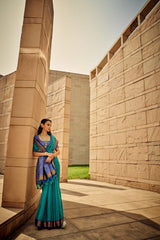 Woman wearing deep green saree with intricate zari work, posing against modern stone architecture under clear sky