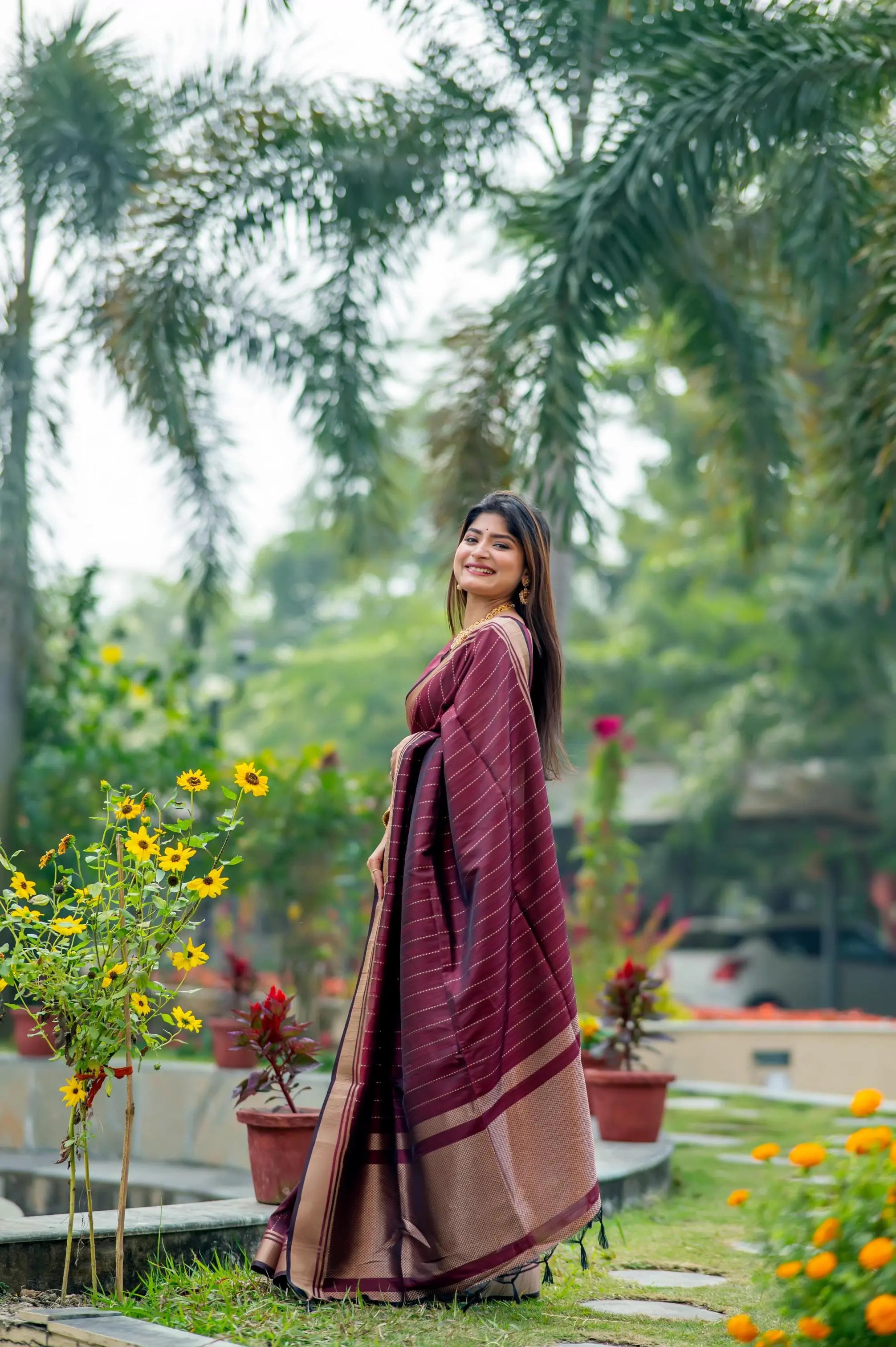 Woman in a deep purple saree with zari work standing in a lush garden with yellow flowers and palm trees in the background
