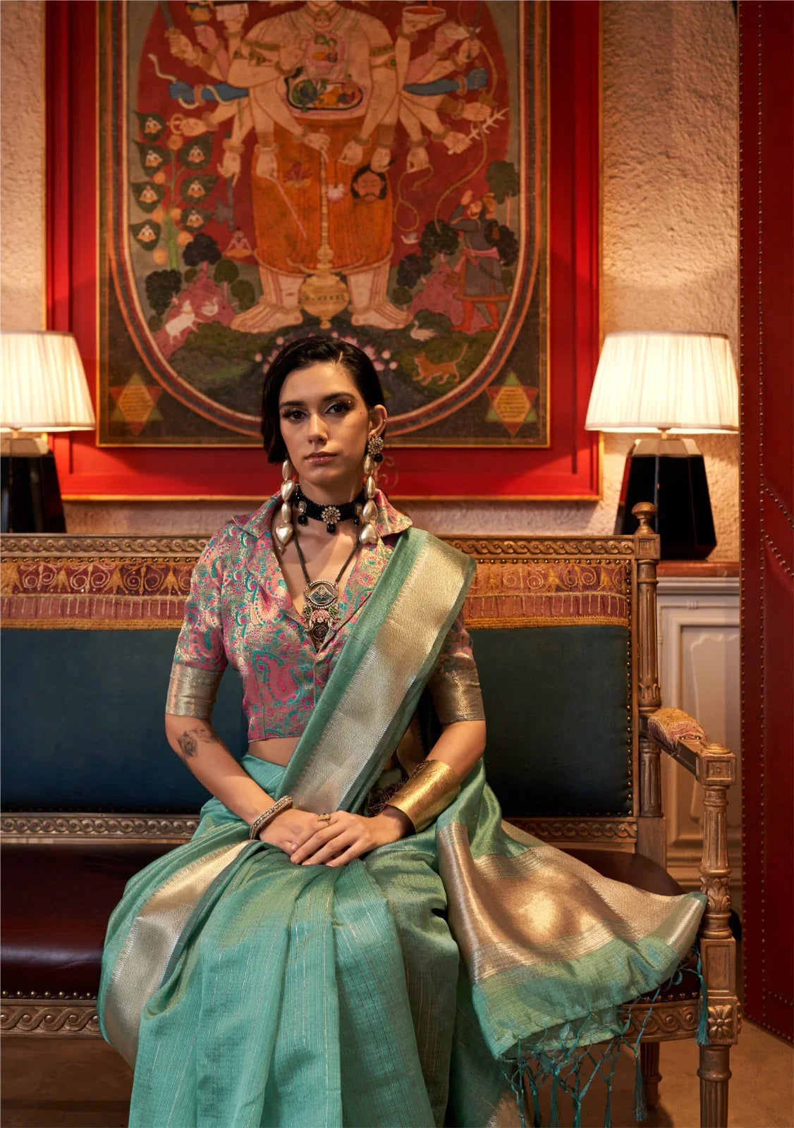 Woman in deep teal zari work saree paired with vibrant embroidered blouse, seated gracefully in a regal ethnic setting