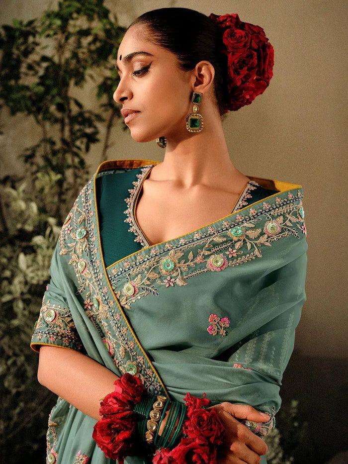 Woman in a dim gray saree with zari work standing near a pond with lotus flowers in a lush garden setting