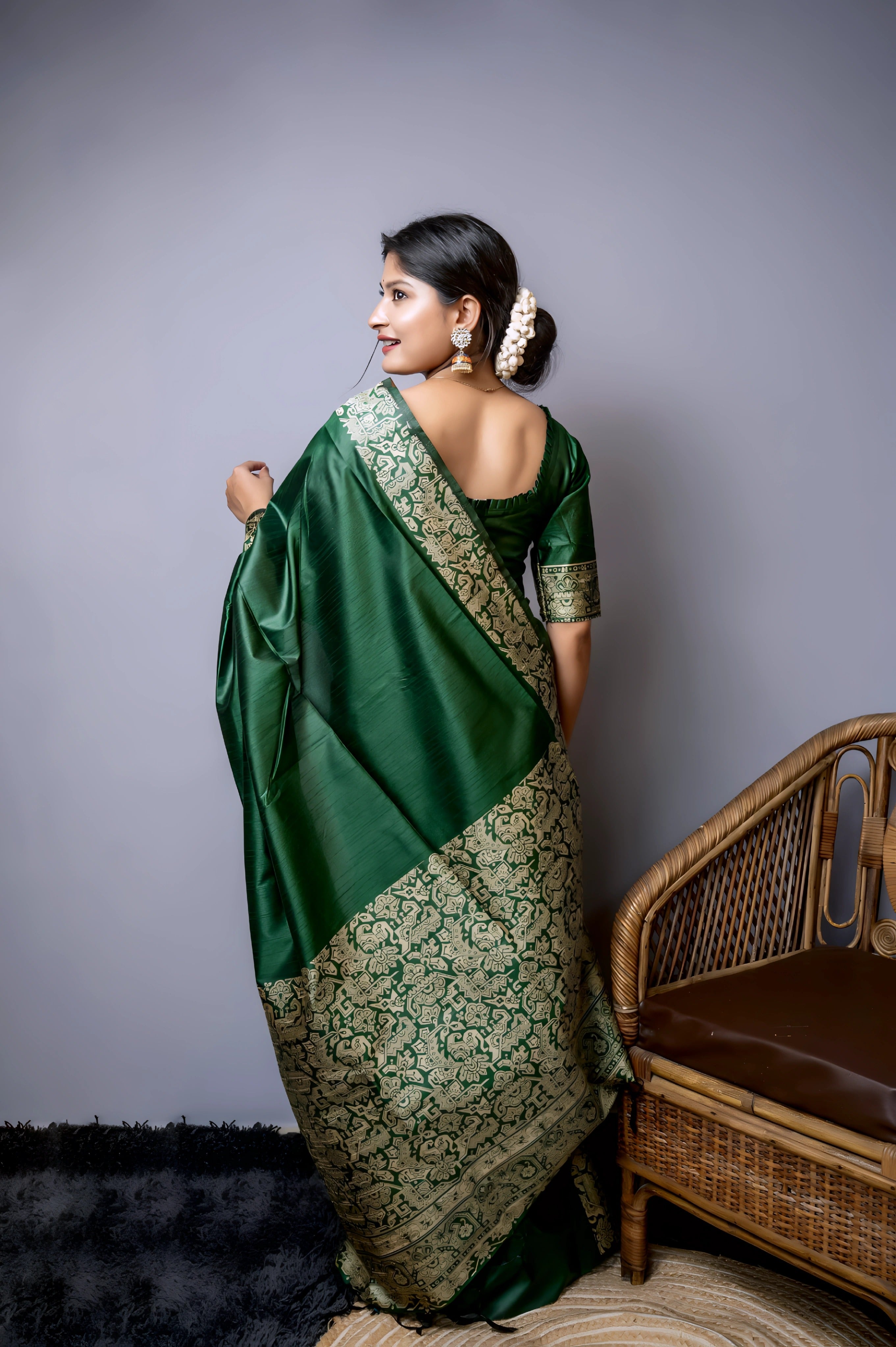 A woman models a luxurious Dark Green Raw Silk Saree. The saree features a wide border and pallu with dense, intricate gold Zari weaving in a Kalamkari-style floral and paisley pattern. She wears a matching short-sleeve green blouse and traditional gold jewelry, including long drop earrings and bangles, with white flowers adorning her hair.