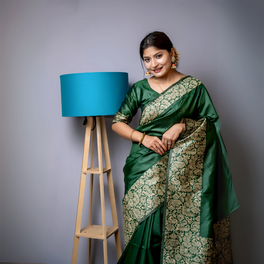 A woman models a luxurious Dark Green Raw Silk Saree. The saree features a wide border and pallu with dense, intricate gold Zari weaving in a Kalamkari-style floral and paisley pattern. She wears a matching short-sleeve green blouse and traditional gold jewelry, including long drop earrings and bangles, with white flowers adorning her hair.