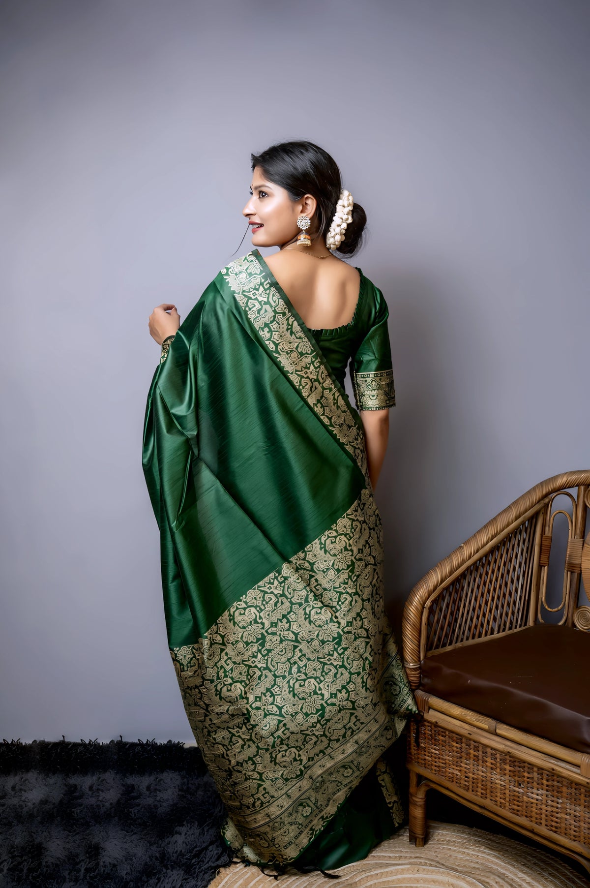 A woman models a luxurious Dark Green Raw Silk Saree. The saree features a wide border and pallu with dense, intricate gold Zari weaving in a Kalamkari-style floral and paisley pattern. She wears a matching short-sleeve green blouse and traditional gold jewelry, including long drop earrings and bangles, with white flowers adorning her hair.