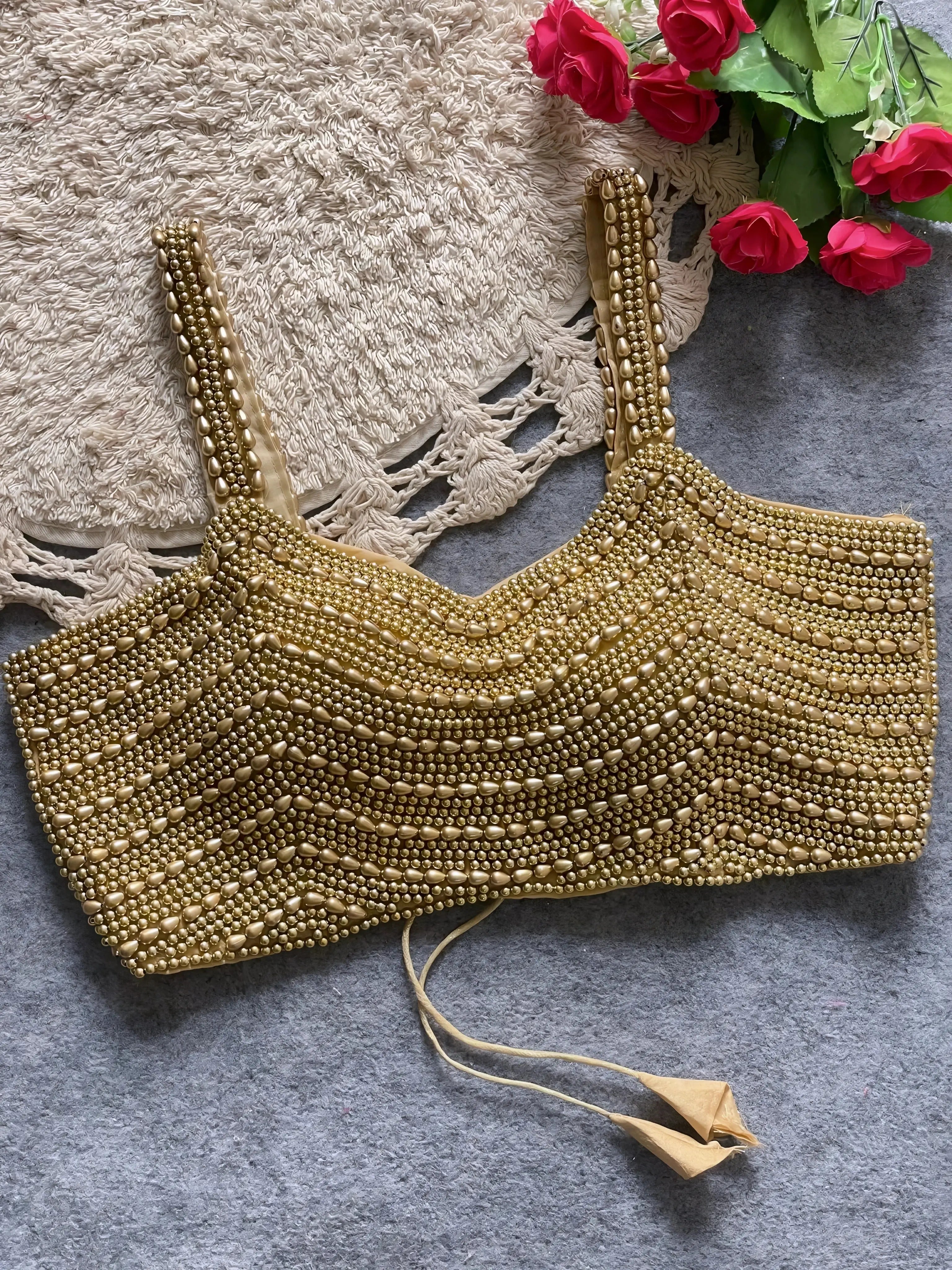 Traditional gold blouse featuring intricate beaded and sequin embellishments in horizontal rows, wide shoulder straps, and a tasseled tie-back detail, displayed flat on a textured grey surface with a braided cream mat and red artificial roses in the corner.