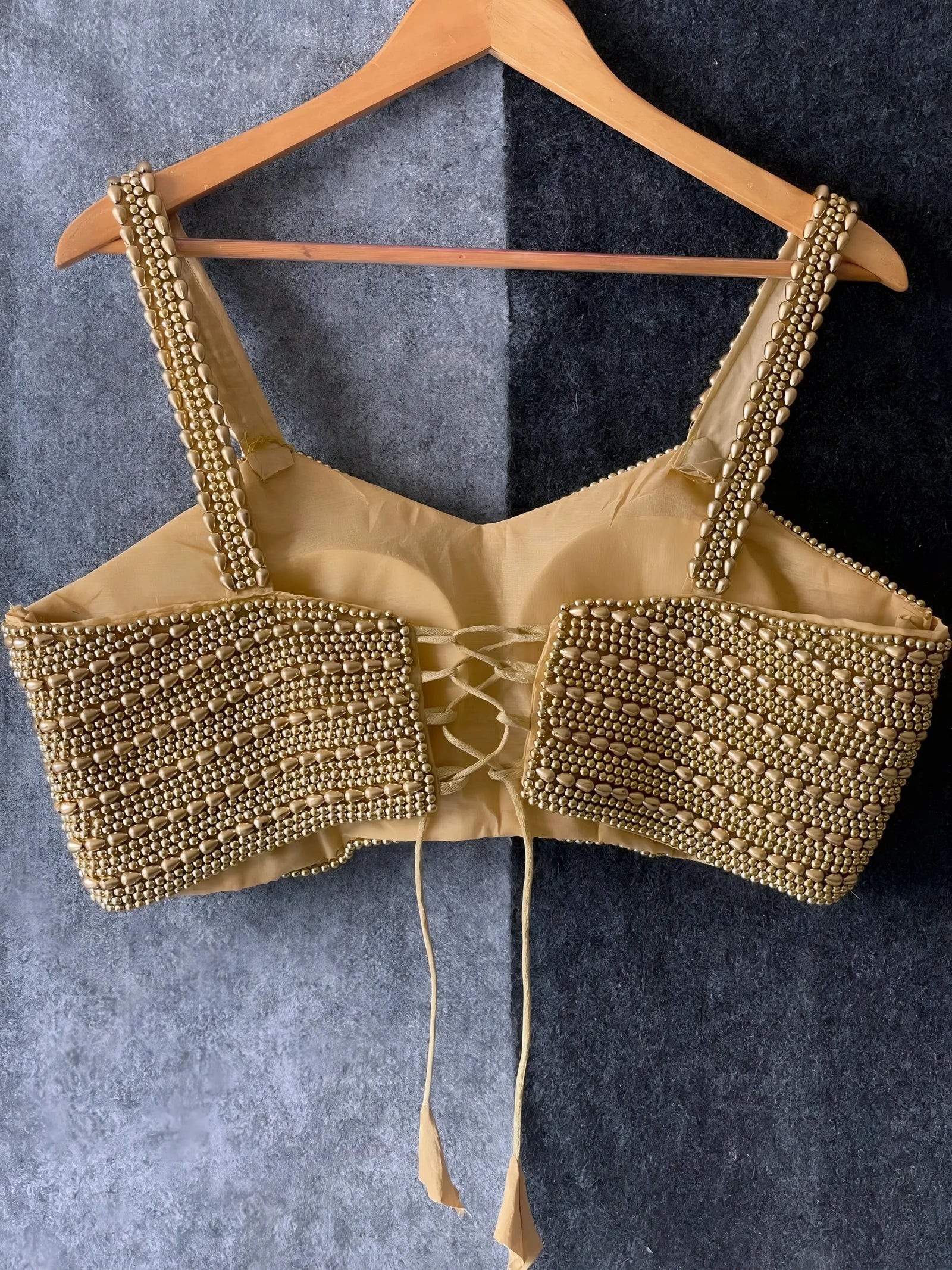 Traditional gold blouse featuring intricate beaded and sequin embellishments in horizontal rows, wide shoulder straps, and a tasseled tie-back detail, displayed flat on a textured grey surface with a braided cream mat and red artificial roses in the corner.