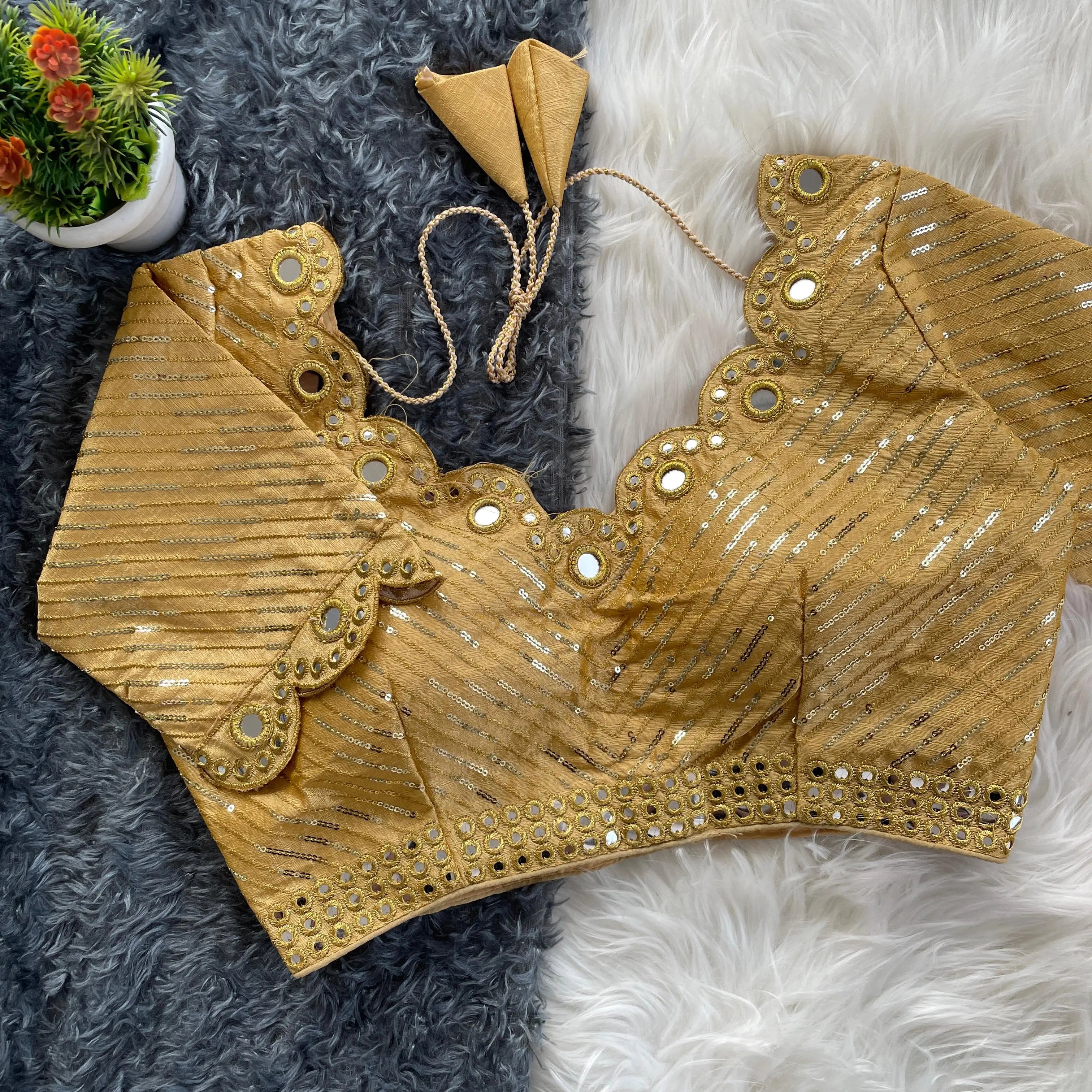 Gold blouse with silver sequin stripes, scalloped neckline embellished with mirror work and decorative borders, displayed on a textured grey and white fur background with a small potted plant nearby.