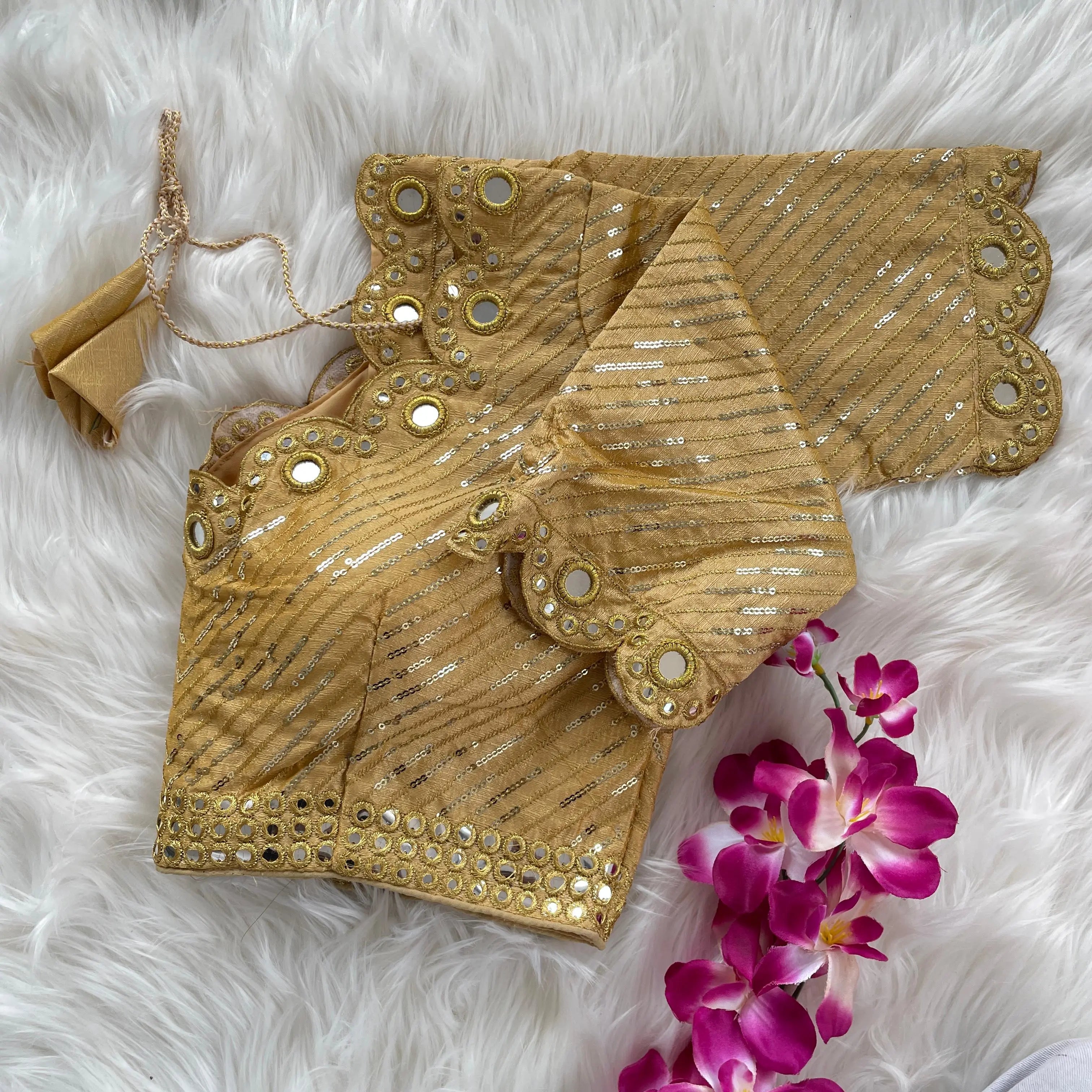 Gold blouse with silver sequin stripes, scalloped neckline embellished with mirror work and decorative borders, displayed on a textured grey and white fur background with a small potted plant nearby.