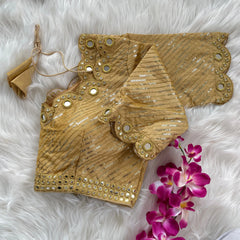 Gold blouse with silver sequin stripes, scalloped neckline embellished with mirror work and decorative borders, displayed on a textured grey and white fur background with a small potted plant nearby.