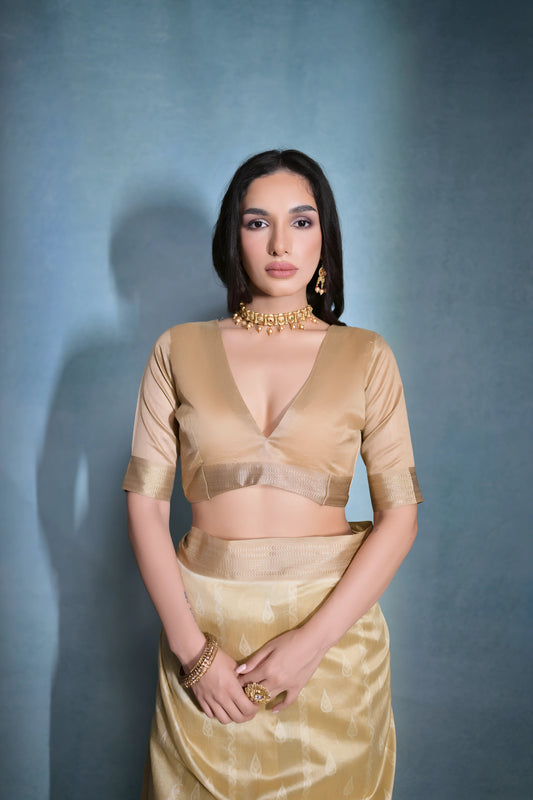 A woman models an elegant Beige/Champagne Soft Raw Silk Saree with a wide metallic gold Zari border. The pallu features distinct horizontal bands of dark brown/black Ikat-style geometric weaving, providing a strong contrast. She wears a matching beige blouse and accessorizes with a gold choker necklace and large gold earrings, posing indoors.
