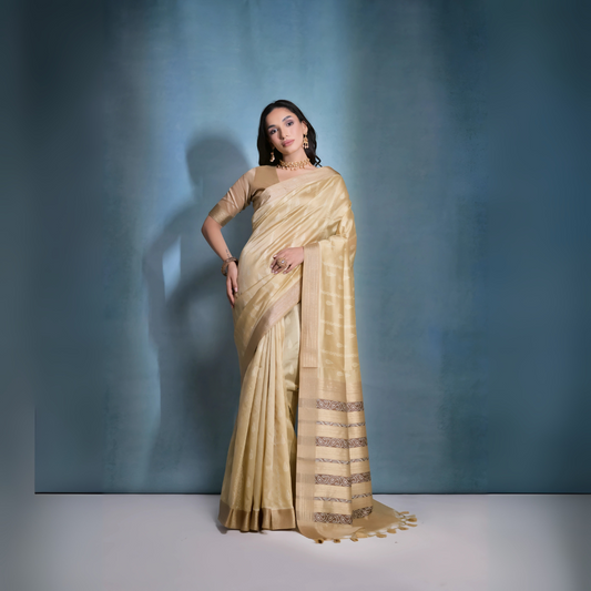 
A woman models an elegant Beige/Champagne Soft Raw Silk Saree with a wide metallic gold Zari border. The pallu features distinct horizontal bands of dark brown/black Ikat-style geometric weaving, providing a strong contrast. She wears a matching beige blouse and accessorizes with a gold choker necklace and large gold earrings, posing indoors.