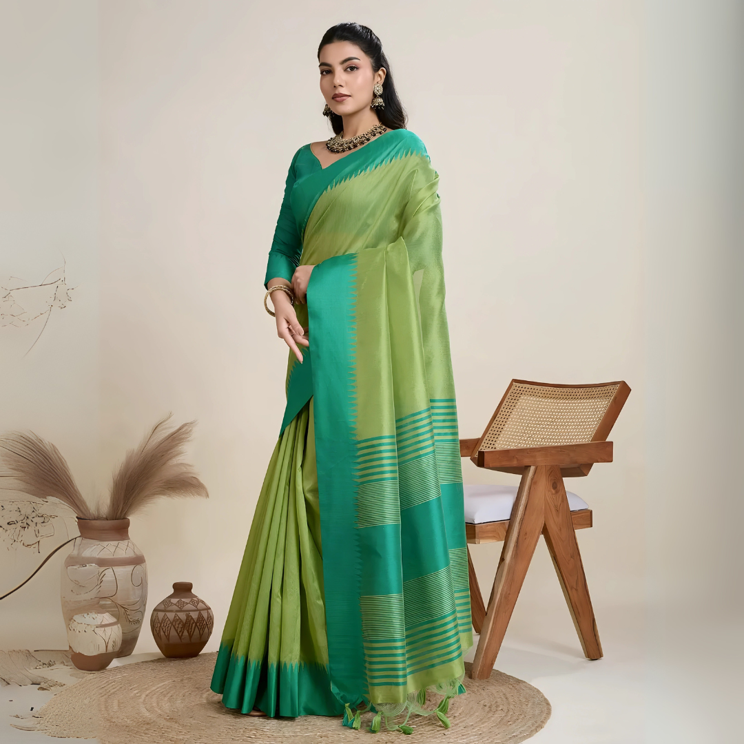 A woman models a striking two-toned Green Raw Silk Saree, transitioning between a light Pista Green and a deep Teal. The pallu contrasts dramatically in a wide band of Teal, featuring horizontal stripe and traditional weaving. She wears a matching short-sleeve teal blouse and accessorizes with a chunky dark gemstone necklace and large gold earrings, posing indoors.